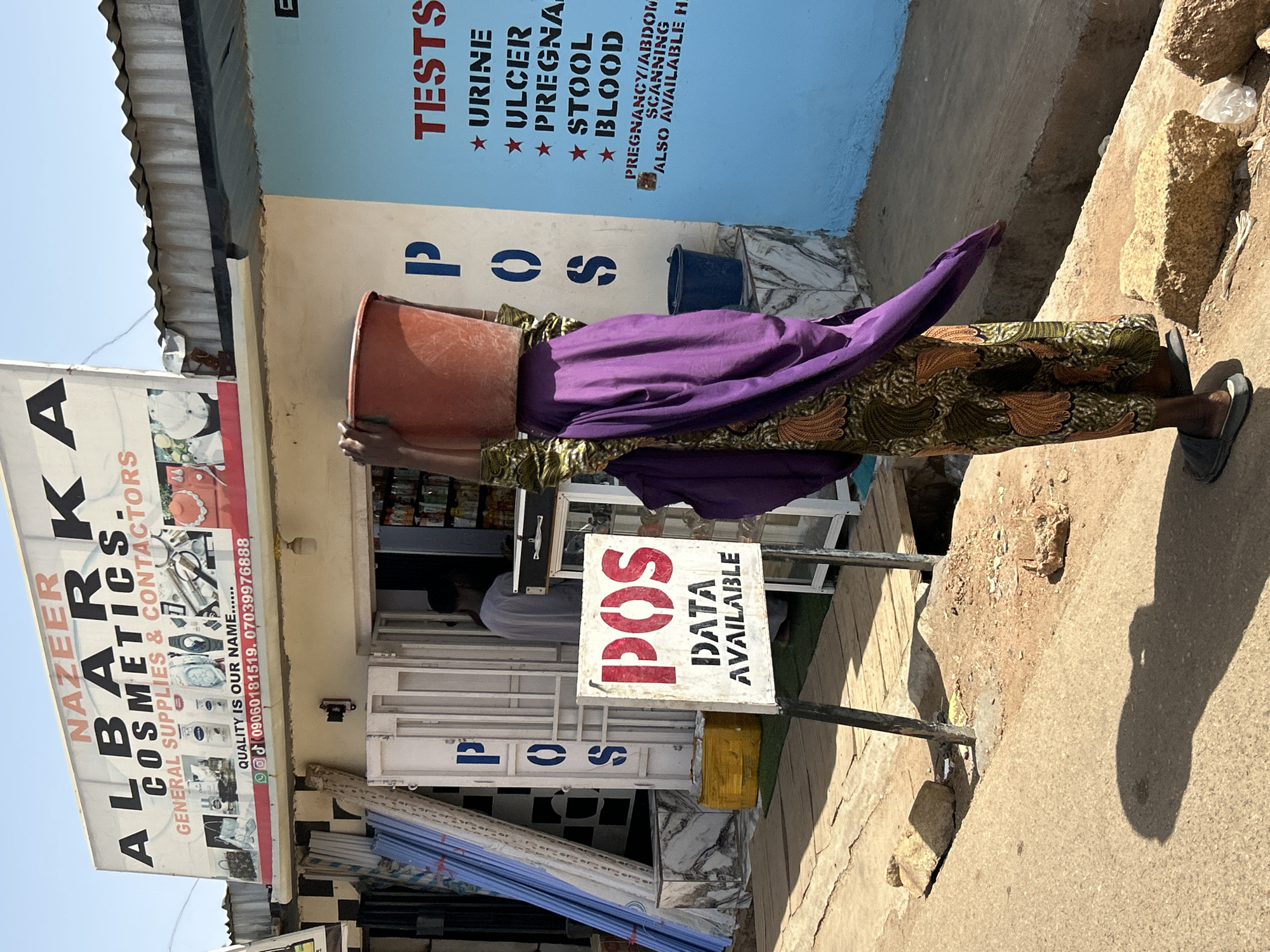 A woman balances a large, orange bucket on her head while walking on a sidewalk in front of a commercial building. She is wearing a patterned skirt, a purple top, and black sandals. The building houses a store called "ALBARKA COSMETICS" and has a sign indicating that medical tests such as urine, ulcer, pregnancy, stool, and blood tests are available. A separate sign advertises "POS DATA AVAILABLE". The scene is located in Jos, Nigeria.