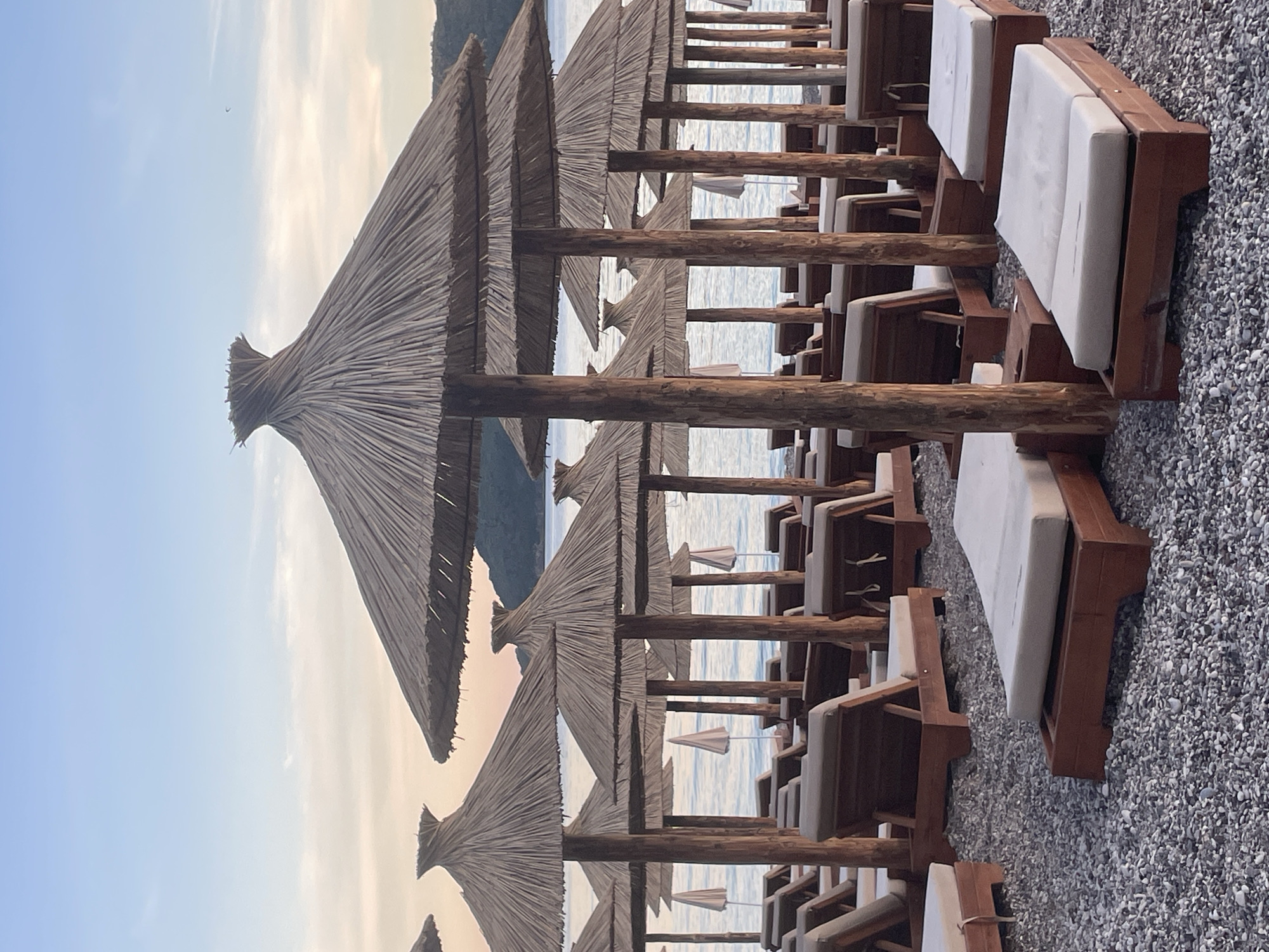 The image depicts a serene beach scene in Budva, Montenegro, with rows of empty lounge chairs and thatched umbrellas on a pebble-covered shore. The water is calm and reflects the soft hues of the sky at dusk. In the background, hills are visible against the horizon. A kite surfer is visible in the distant sky. The scene evokes a sense of tranquility and anticipation for the evening to come.
