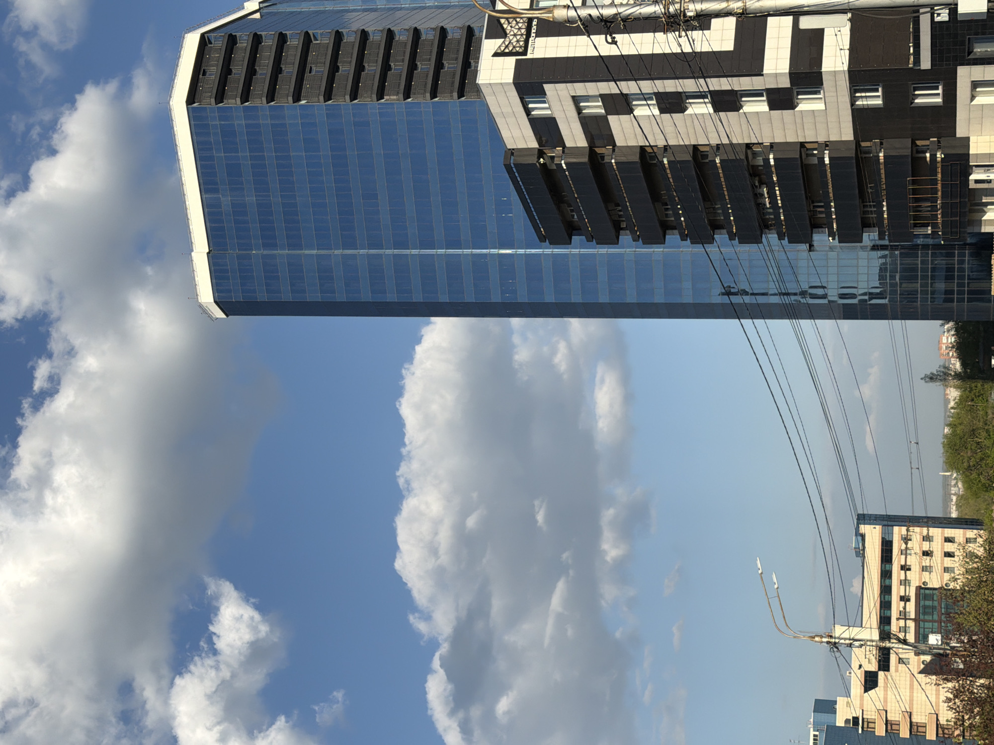 The image depicts a cityscape of Krasnoyarsk, Russia, featuring a modern skyscraper with a reflective glass facade and a section with alternating black and white bands. The skyscraper is positioned on the left, dominating the frame. A cluster of white clouds is visible behind it, contrasting with the blue sky. Power lines are strung across the sky, running diagonally through the image. In the background, another building is visible, with a lighter, beige and white exterior. Trees and foliage can be seen in the distance, indicating a green space or park area. The perspective is tilted, giving an upward view of the buildings.
