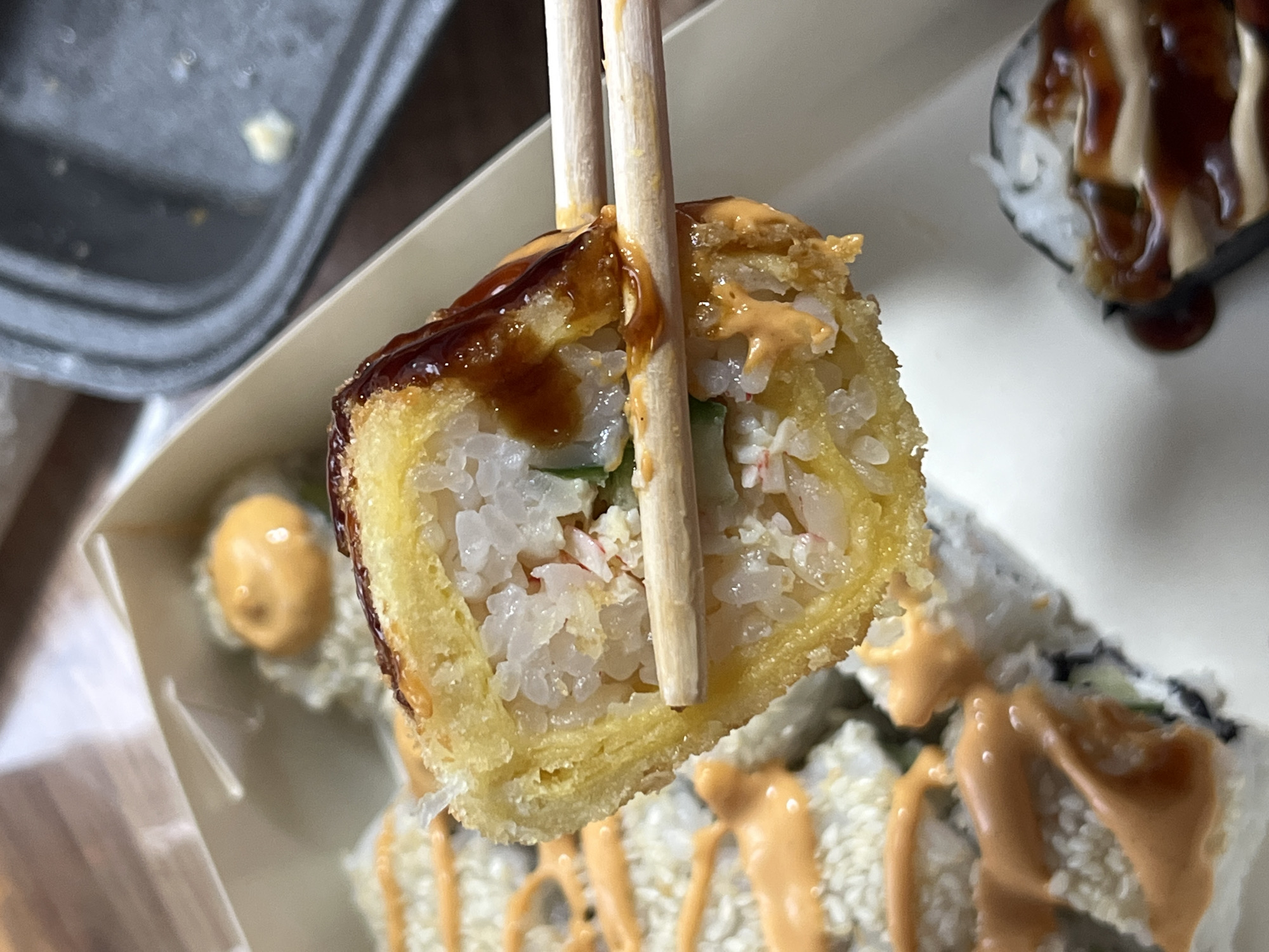 A close-up shot captures a piece of sushi being held by two chopsticks. The sushi roll is deep-fried, with a golden-brown exterior and a visible filling of rice, what appears to be imitation crab meat, and possibly cucumber. It is drizzled with a dark, glossy sauce, likely teriyaki or eel sauce, and a creamy orange sauce, possibly spicy mayonnaise. The sushi is presented in a light-colored cardboard container, with other sushi rolls visible in the background, also decorated with the orange sauce and sesame seeds. To the left, a dark plastic container, possibly part of the takeout packaging, is partially visible. The lighting suggests an indoor setting, likely a restaurant or home. The overall impression is one of a prepared meal, ready to be eaten. There is no visible text in the image. The setting is not specified beyond being a meal, and no temporal or emotional cues are apparent. Given the context provided, the location is Nizhny Tagil, Russia, though this is not visually discernible from the image itself.
