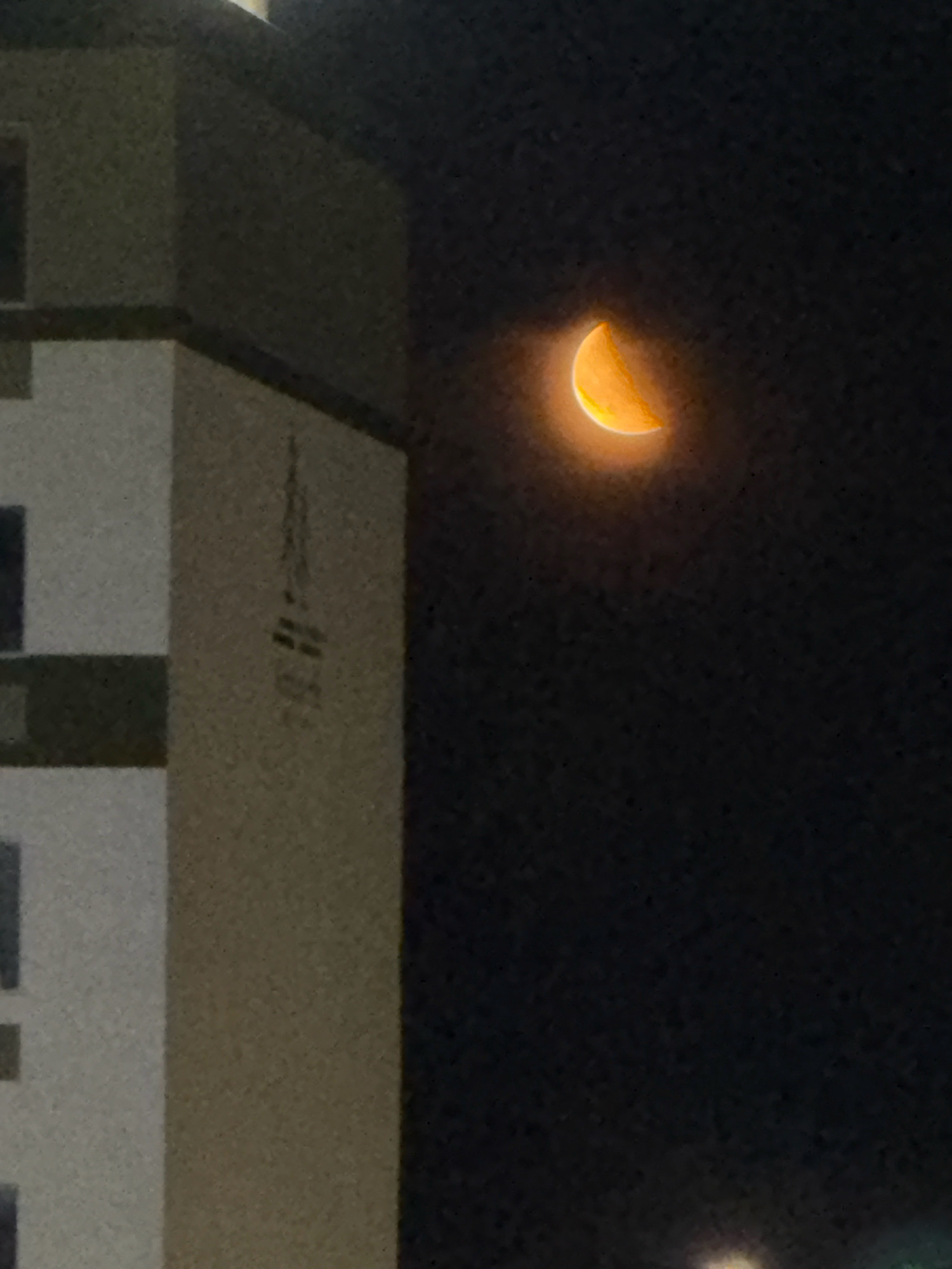 A crescent moon glows with an orange hue in a dark night sky above the city of Astana, Kazakhstan. To the left of the frame, the corner of a light-colored building with dark windows is visible, and further to the left, a darker section of the building is present. The building's facade has some faint markings that are difficult to discern. The overall impression is a dimly lit urban environment illuminated by the celestial body.