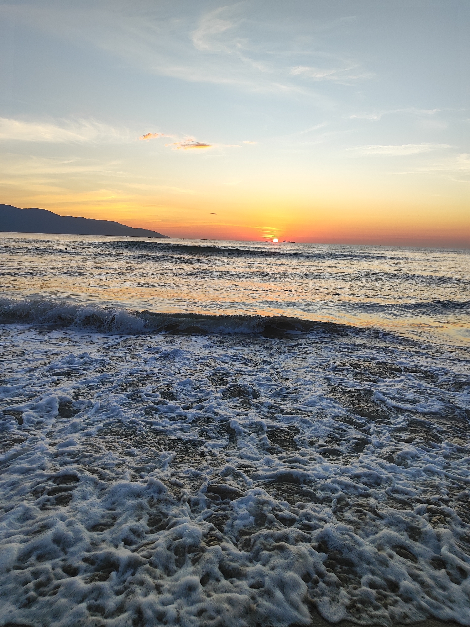 The image depicts a serene beach scene at sunrise in Da Nang, Vietnam. The sky is a gradient of soft blues and oranges, with the sun just beginning to crest the horizon, casting a warm glow on the water. In the distance, a silhouette of mountains lines the shore. The foreground is dominated by the gentle ebb and flow of the ocean, with waves creating a frothy white foam that washes onto the sandy beach. There are no visible people in the image, and only a few distant boats can be spotted on the water. The overall impression is one of tranquility and natural beauty, characteristic of Da Nang's coastline during the early morning hours.