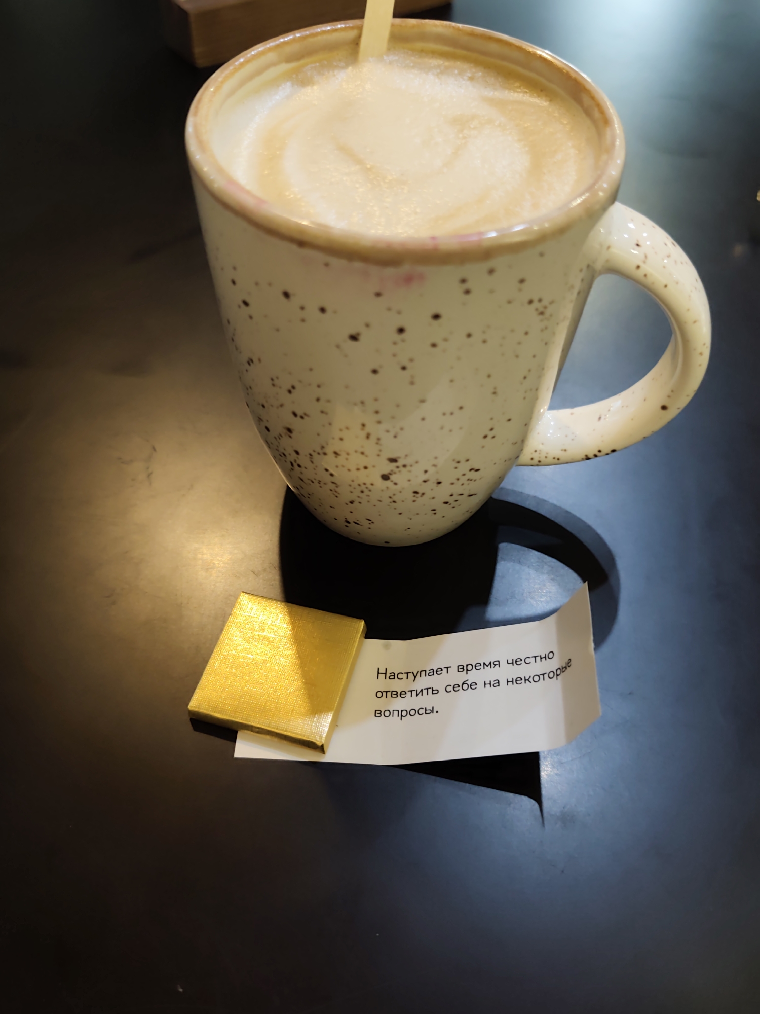 A close-up, high-angle shot shows a beige speckled mug with a creamy beverage and a yellow fortune cookie wrapper on a dark, textured surface, likely a table. A golden, rectangular object, possibly a small box or foil wrapper, sits to the left of the fortune cookie paper. The fortune cookie paper has text in Russian that translates to "The time has come to honestly answer some questions." The lighting suggests an indoor setting, with a warm light source creating a shadow from the mug. The background is dark and slightly out of focus, with a hint of a wooden object to the upper left. The overall scene evokes a casual, possibly cafe or home, environment. There is no indication of the city of Sochi, Russia.