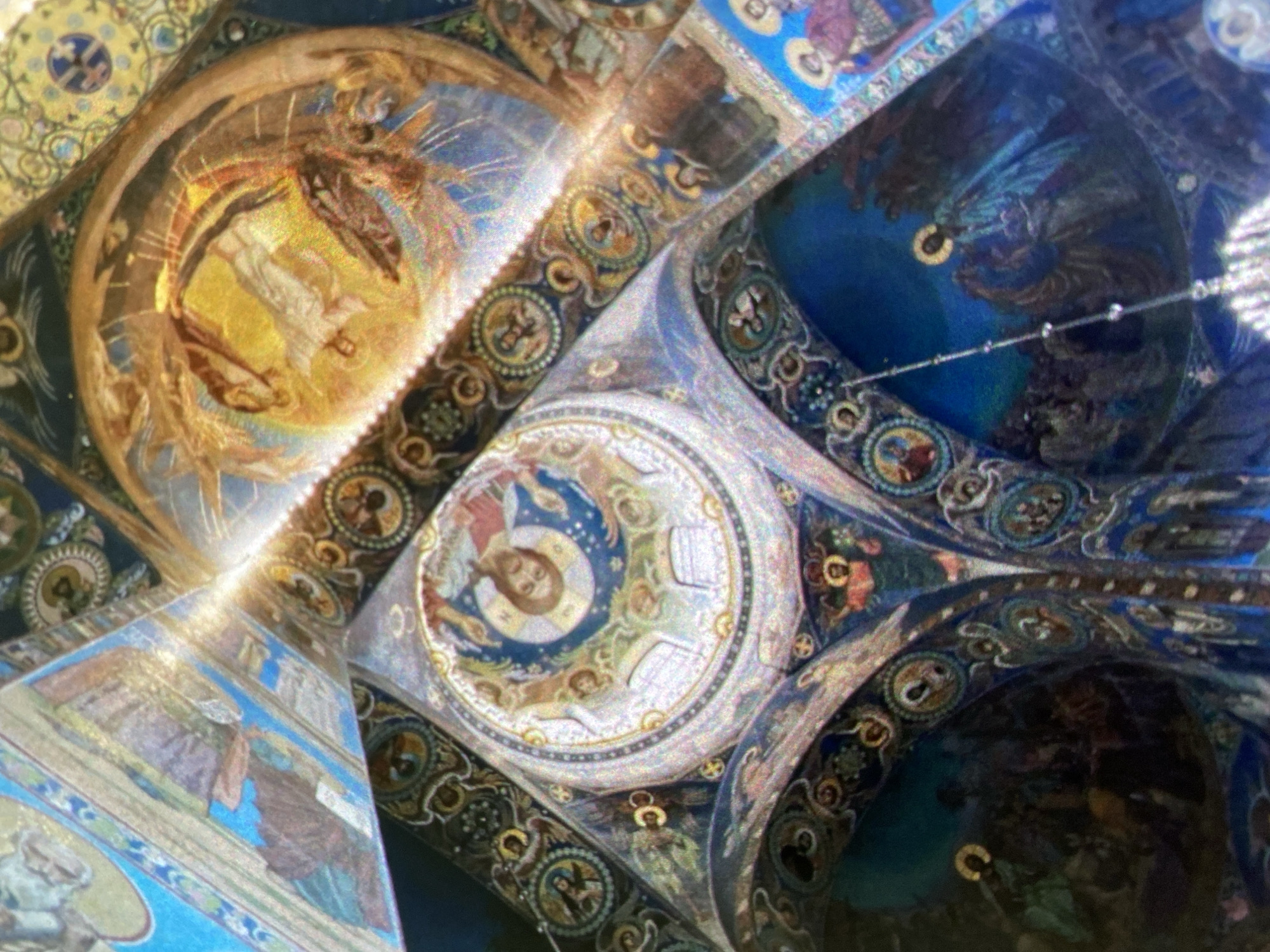 This image shows the interior of the Church of the Savior on Spilled Blood in Saint Petersburg, Russia, focusing on the mosaic-covered ceiling. The picture is taken from below, looking upwards at the intricate designs. The primary subjects are the detailed mosaic depictions of biblical figures and scenes, characteristic of Russian Orthodox church architecture. Visible are scenes within golden circles, surrounded by detailed ornamentation in various colors, including blue, gold, and earth tones. The scene features a section with a golden haloed figure encircled with a rainbow-hued surrounding and other depictions of saints with books and halos. The environment consists of the ornate ceiling, showcasing the impressive craftsmanship of the church's interior.