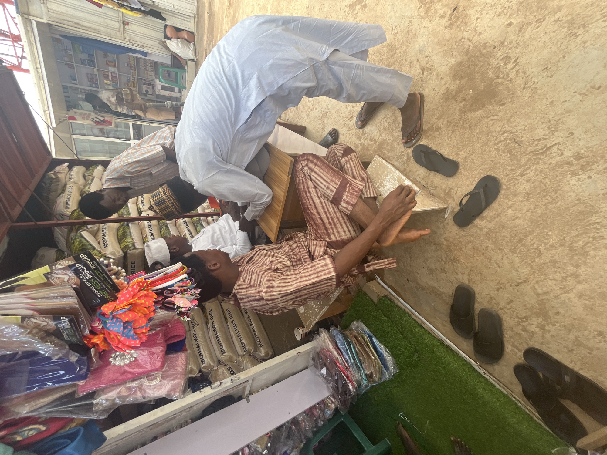 The image depicts an outdoor marketplace scene in Abuja, Nigeria. In the foreground, a man wearing a light blue traditional robe and trousers is leaning over a wooden table, interacting with another man. The second man, seated on a low bench, wears a striped brown and white traditional outfit. Behind them, stacks of "MOHALKOS RICE" bags are visible. To the left, various items are displayed, including packaged goods and colorful hair accessories hanging from the ceiling. The ground is a mixture of concrete and artificial turf. The overall impression is a bustling commercial environment.