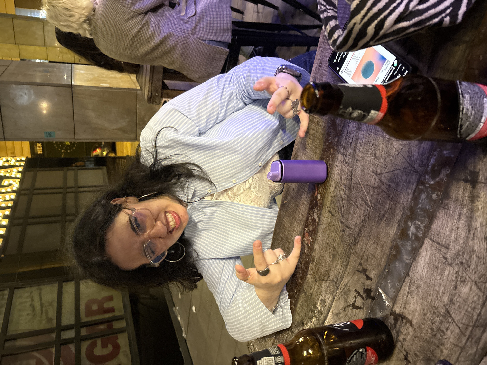 A woman with dark hair and glasses sits at a weathered wooden table in what appears to be a bar or restaurant. She is wearing a light blue striped shirt over a white lace top. She's making a rock-on gesture with both hands. Two open bottles of Alhambra beer sit on the table, along with a purple electronic device. There's a cell phone with the screen visible, a zebra print handbag and another person sitting at the table out of focus. Through a window, the signs of "Gar" are visible, suggesting the location in Cordoba, Spain, near the restaurant called El Garum 2.1.