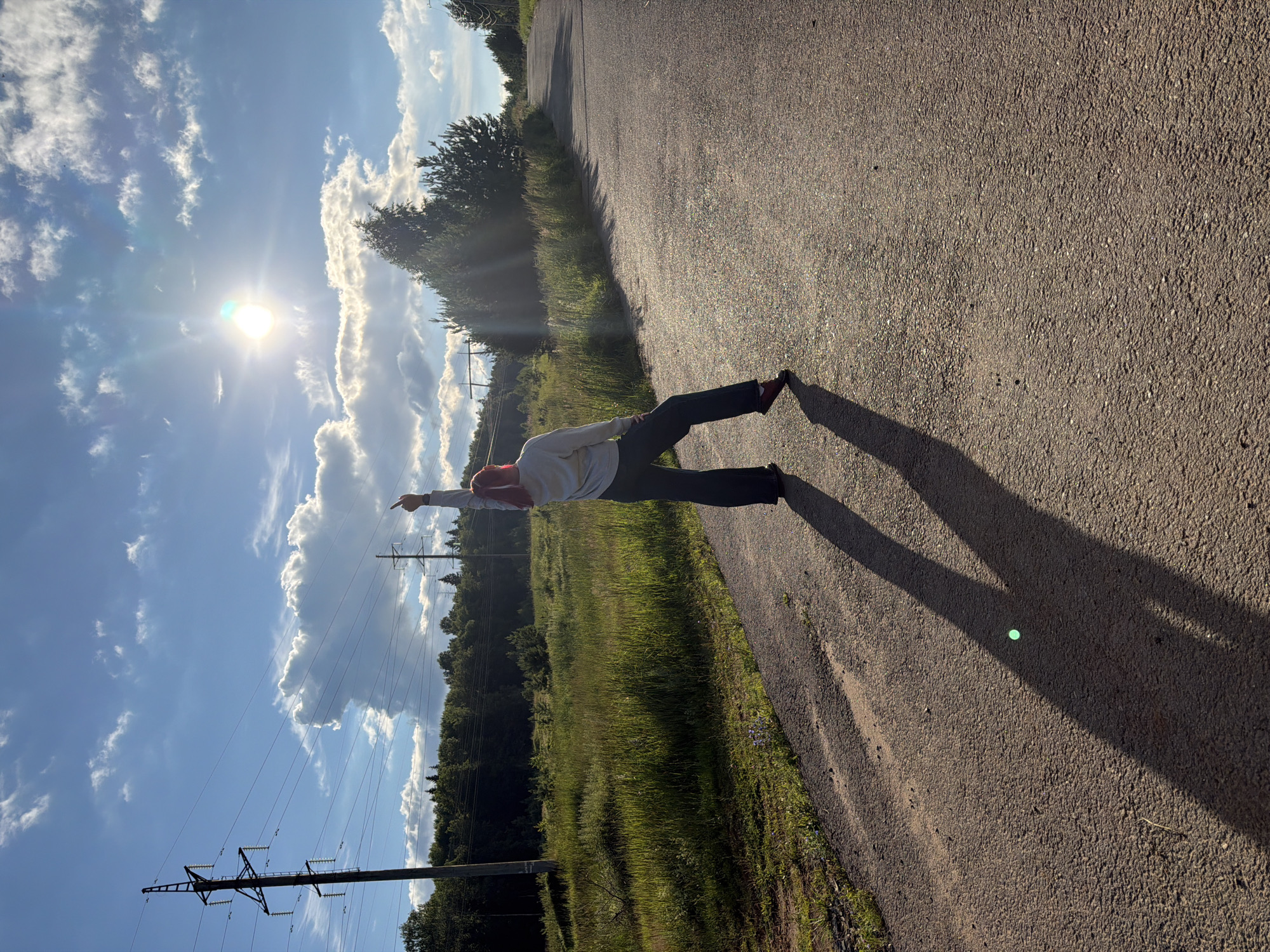 A person with bright red hair, wearing a light-colored hoodie and dark pants, stands on an asphalt road with their right arm raised, index finger pointing upwards. The sun is shining brightly in a blue sky with scattered clouds. The person is casting a long shadow on the road. To the left of the road, there is a grassy field with trees in the background and a power line pole. The road curves to the right and recedes into the distance, bordered by trees. The scene appears to be a rural or suburban area. The location is identified as Chaikovsky, Russia.