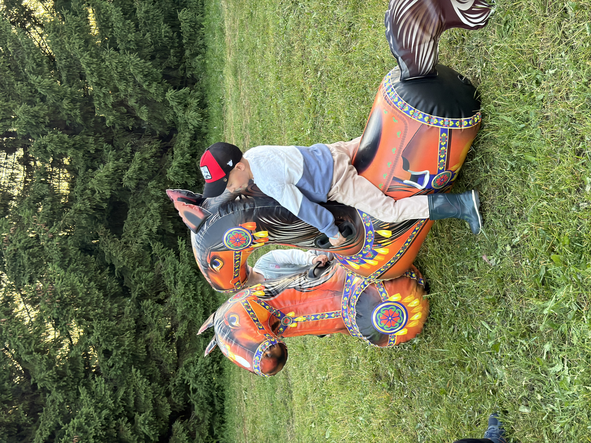 An adult male is seated on an inflatable horse in a grassy outdoor area, with a dense evergreen forest in the background. He is wearing a white and blue long-sleeved shirt, tan pants, and dark green rain boots. He is also wearing a red and black baseball cap. The inflatable horse is orange and black with decorative patterns. Another inflatable horse is positioned next to it. A child, partially obscured, is behind the child on the horse, holding onto the reins. The sun is setting, casting a warm glow through the trees.