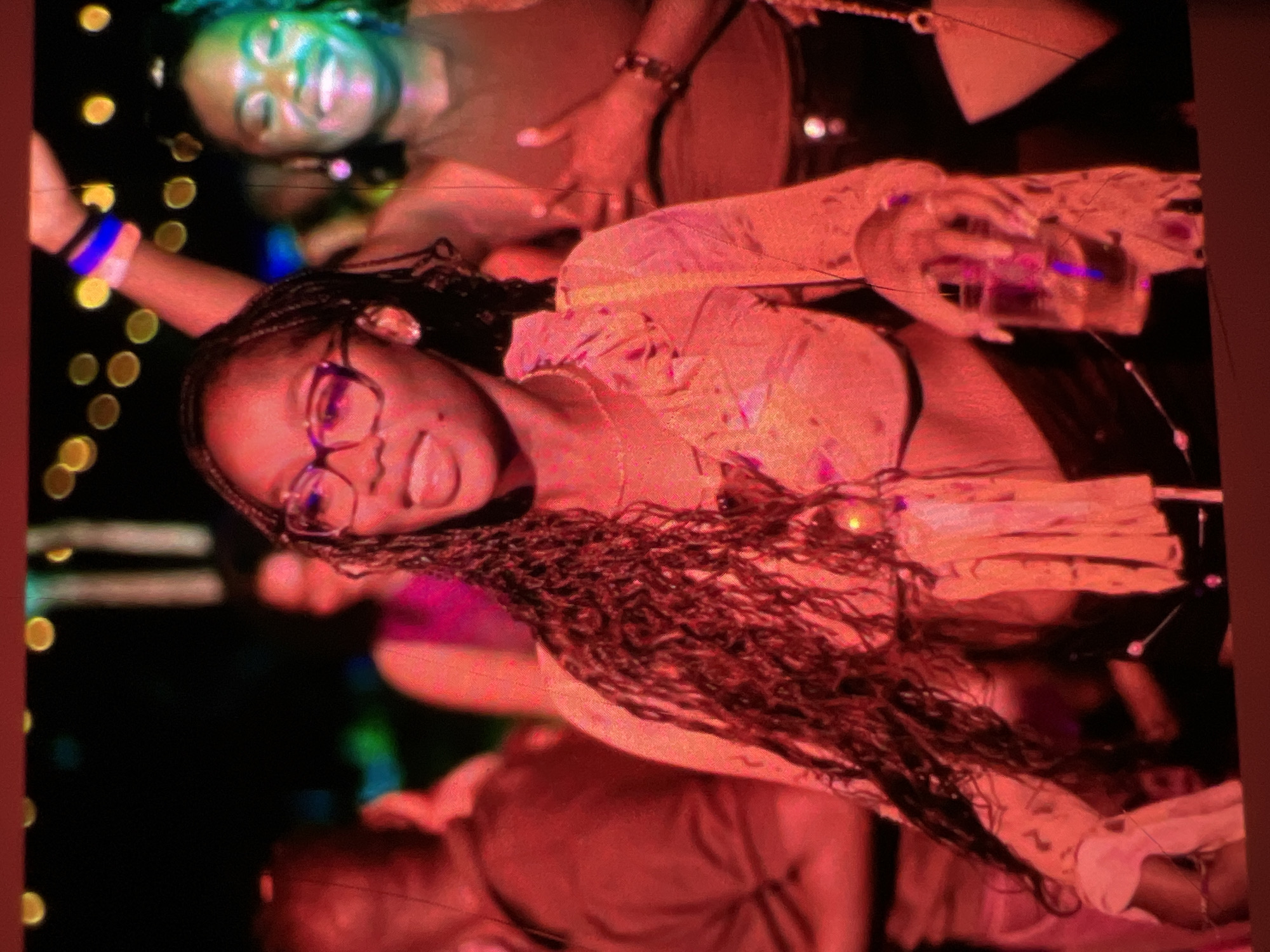 A medium shot captures a young Black woman with long, braided hair and glasses, wearing a light-colored, floral-patterned crop top. She is holding a clear cup with a beverage in her right hand and is looking to her left. Behind her, another woman with closed eyes and a wide smile has her left arm raised, adorned with two blue and purple wristbands. The scene appears to be outdoors at night, possibly at a party or concert, with blurred figures in the background and festive lights overhead. The overall lighting is dim and colorful, with dominant red and orange tones. While the image itself does not provide explicit visual cues to pinpoint its location to Lagos, Nigeria, the context of vibrant nightlife and social gatherings is consistent with urban environments.