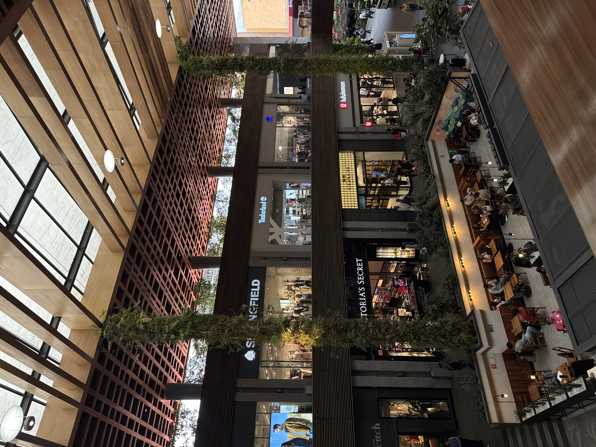 This photograph shows an indoor view of a commercial space. The photo is taken at an upward angle, capturing several floors of a shopping mall or similar commercial structure.
Retail storefronts with visible signage like “Timberland,” "SPRINGFIELD," “Victoria’s Secret,” “Lululemon,” and “Abercrombie and Fitch” can be seen, suggesting the location is a shopping mall. Some stores are active with shoppers inside.
On the right side of the frame, an open-air seating area with tables and chairs is visible, populated with people sitting and possibly dining. Decorative vegetation is interspersed among the architectural elements, particularly along the vertical columns. The image shows a mix of architectural styles, including wood lattices, and modern building materials.
Based on the businesses featured, the architectural style and decor, and the presence of certain retail chains that are known to operate there, the location can be identified as Parque Puebla Shopping Mall in Puebla, Mexico.