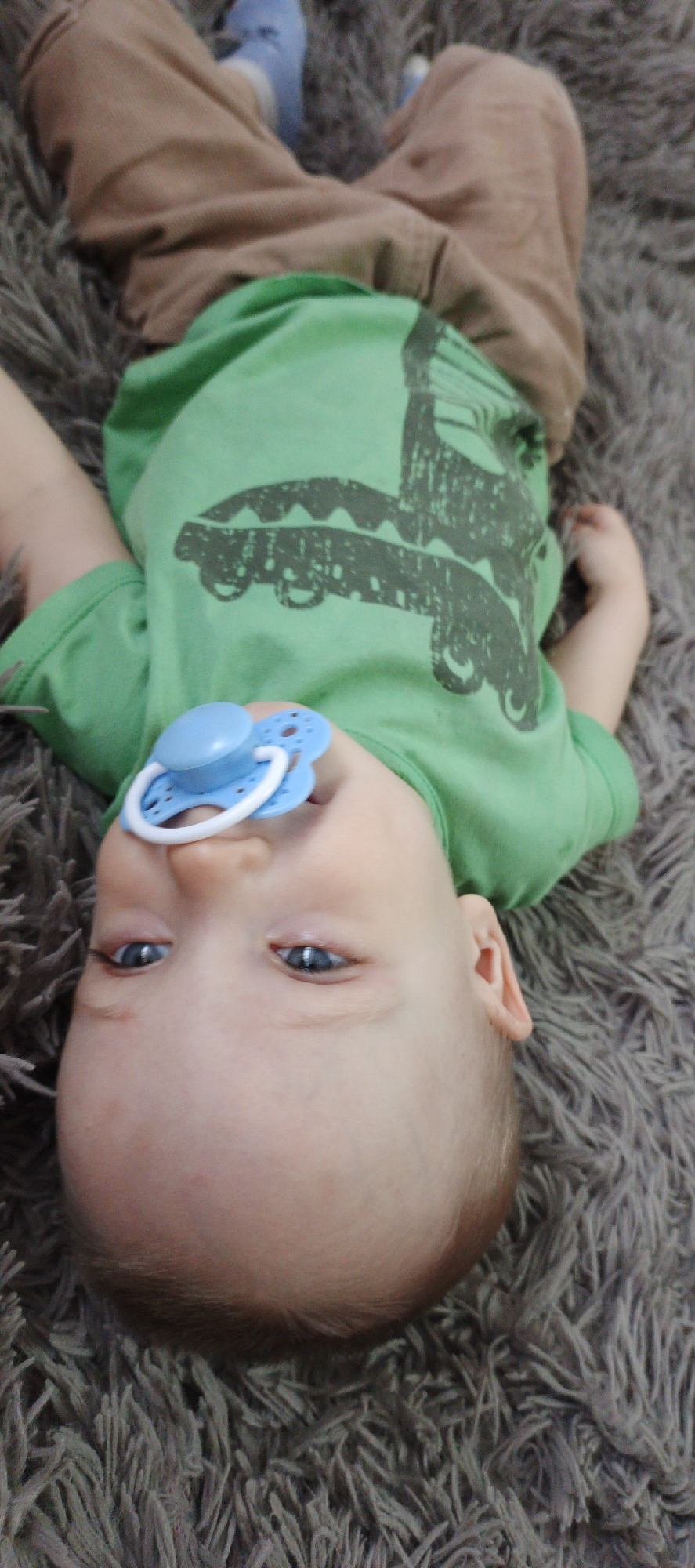 A baby lies on a gray, shaggy rug. The baby wears brown pants and a green t-shirt with a graphic of a vehicle on it. The baby has blue eyes and fair skin, and is using a blue pacifier. The scene appears to be indoors. There is no evidence to suggest that this image was taken in Chaikovsky, Russia.
