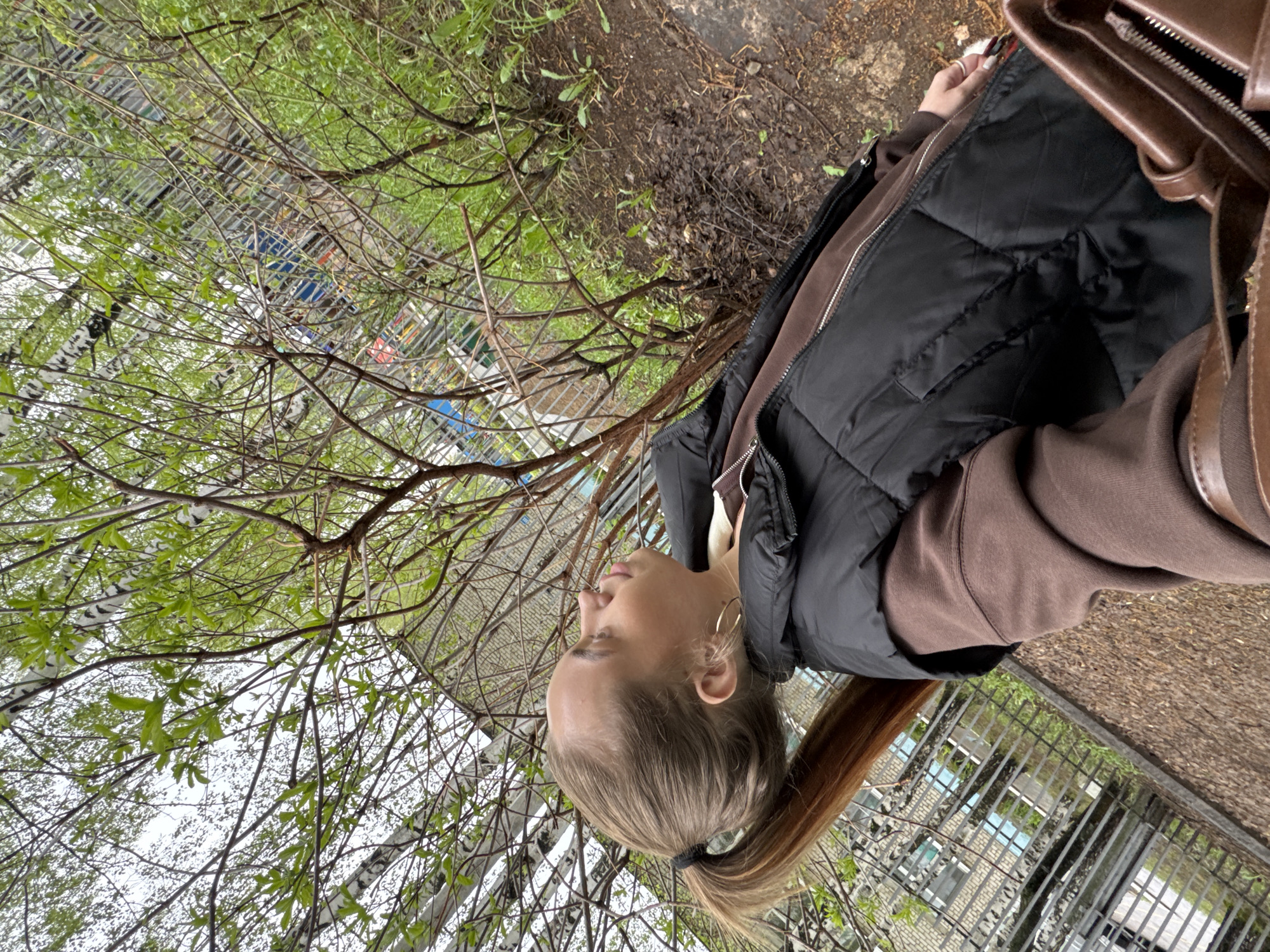 A woman with light brown hair pulled back in a ponytail stands outdoors near a birch tree with new leaves in a cityscape, implied through a fence in the background. She is wearing a brown hoodie and a black puffer vest. A brown handbag is over her shoulder. The location is likely Chaikovsky, Russia, based on request, although there is no evidence to substantiate this within the image. The ground is either dirt or paved with gravel. The weather appears overcast.