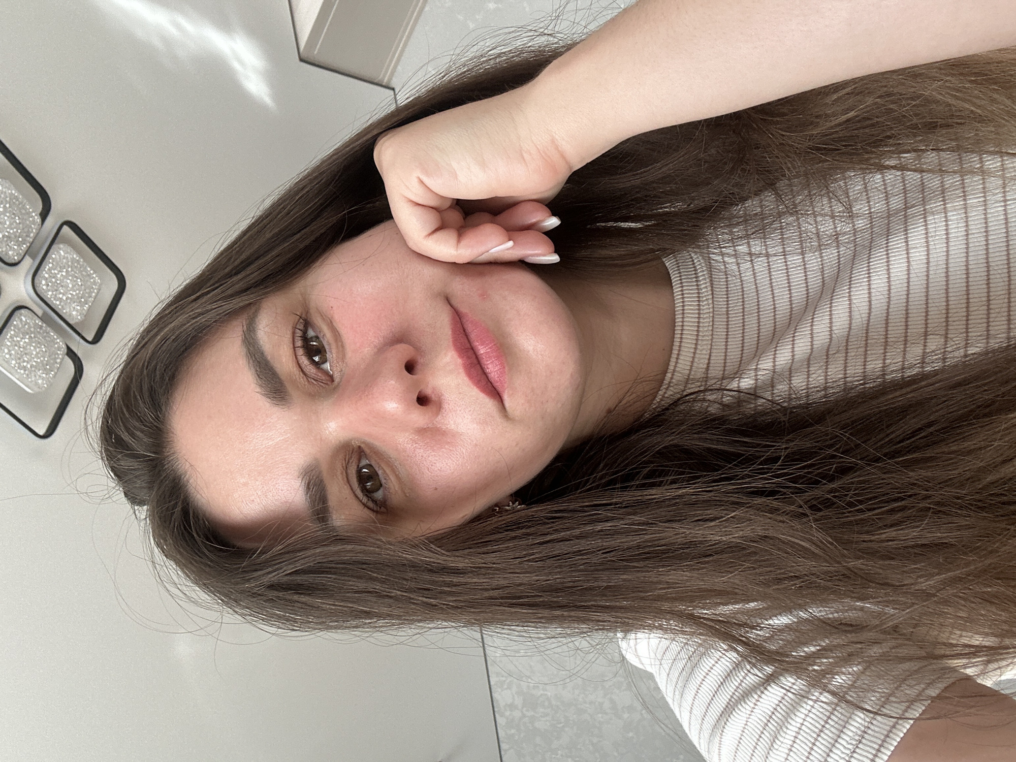 The media file is a selfie of a young woman with long, brown hair. She is wearing a light-colored, striped t-shirt. Her right hand is resting on her cheek, and her fingers are curled. She is looking towards the camera with a neutral expression. The background is a light-colored wall with some decorative lighting fixtures visible on the left side. The lighting suggests it is daytime, possibly indoors. The overall impression is that of a casual, personal photograph. The image appears to be taken in Chaikovsky, Russia, based on the provided location context. No text is visible in the image.