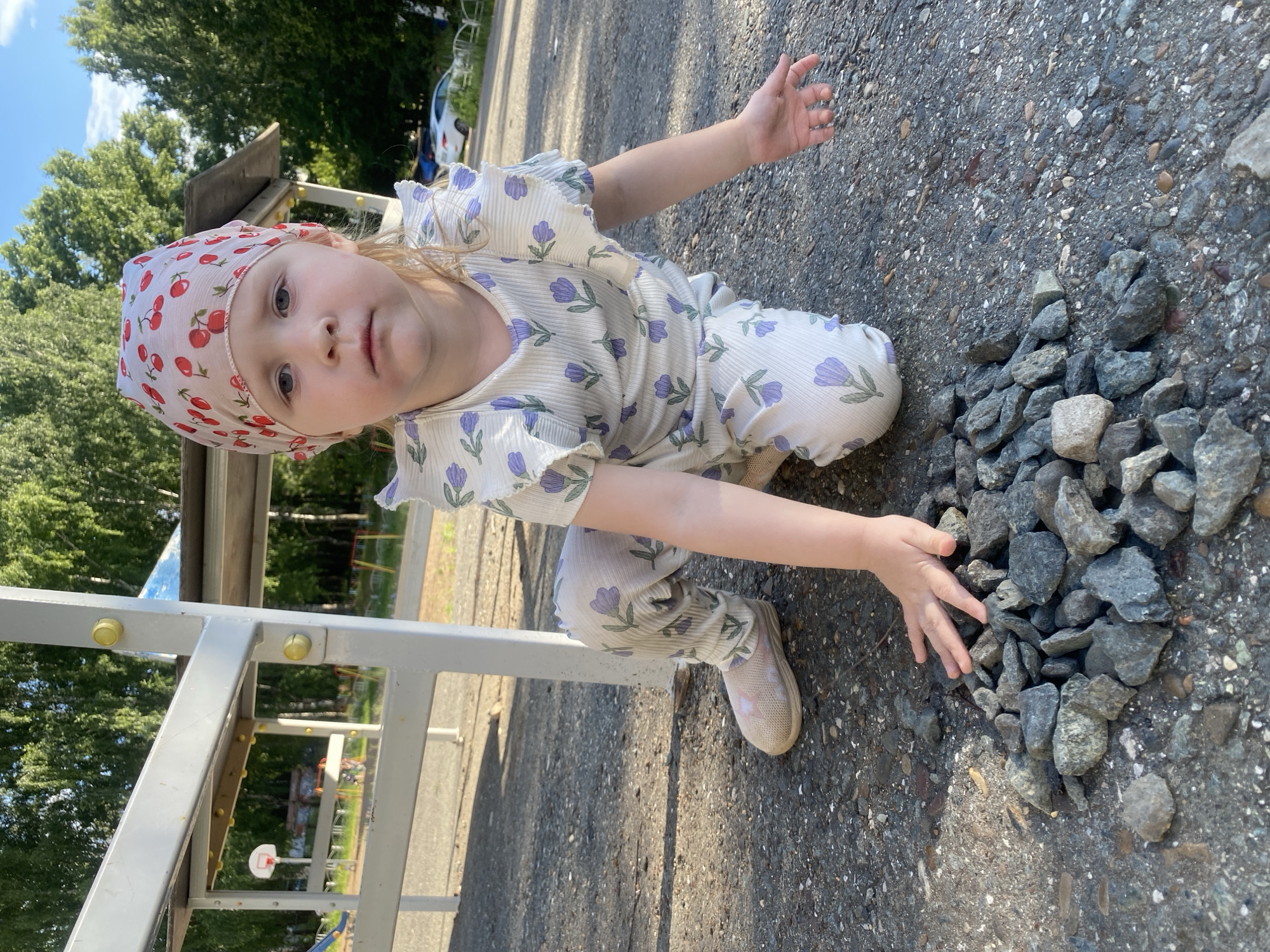 A young girl with blonde hair is kneeling on a gravel surface. She is wearing a white outfit with a pattern of purple flowers and a bandana with red cherries. The girl is looking at the camera with a neutral expression. Her left hand is raised, and her right hand is reaching towards a pile of rocks. The scene appears to be outdoors, with trees and a playground in the background. The lighting suggests it is daytime, likely sunny. There is no visible text in the image.