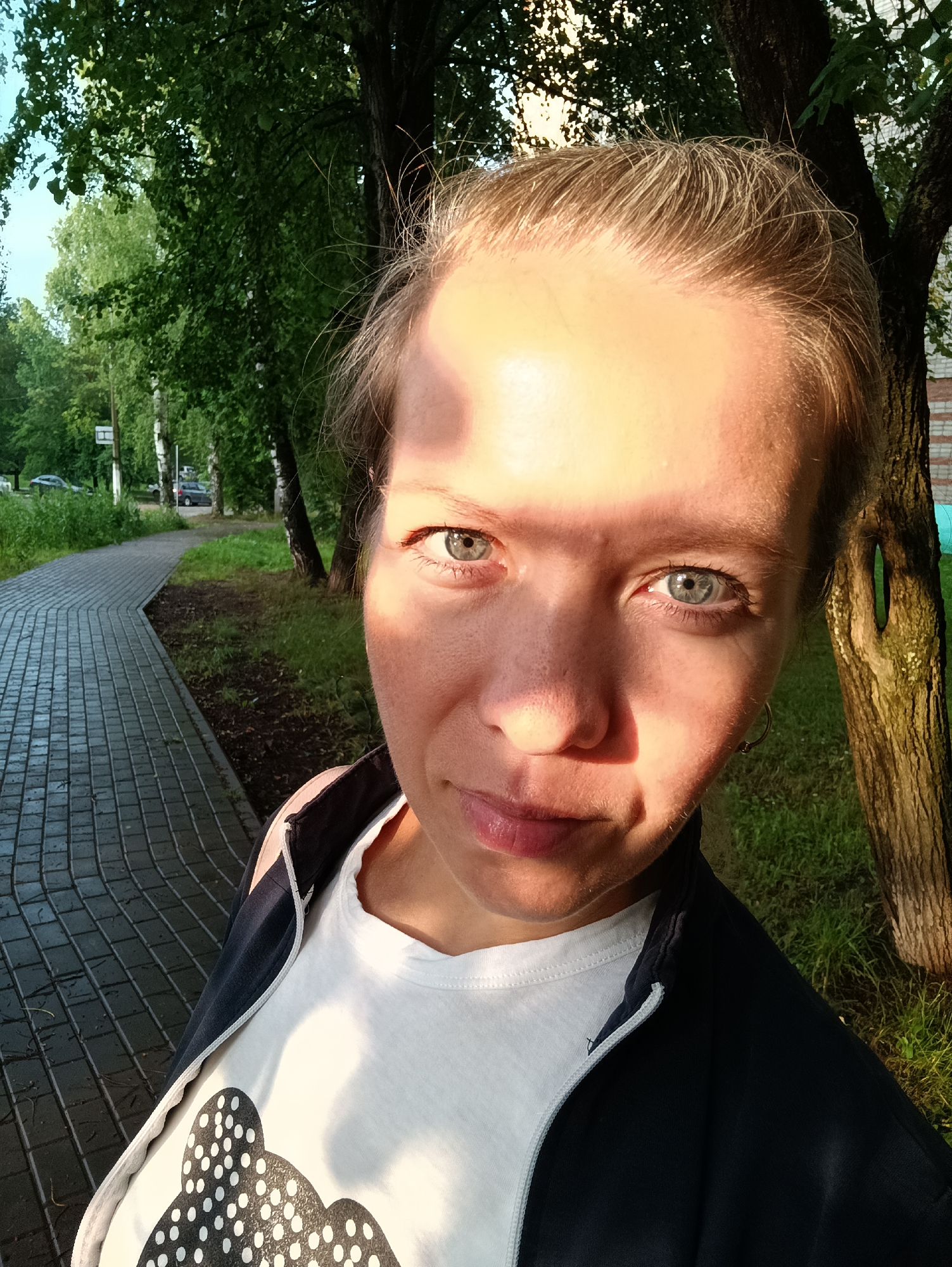 A close-up, selfie-style photograph captures a young woman with blonde hair pulled back, looking directly at the camera. She is positioned to the right of the frame, with her face illuminated by the warm glow of sunlight, casting a shadow across her forehead. Her eyes are a striking blue-grey, and she has a subtle smile. She is wearing a white t-shirt with a black polka-dot pattern on it, and a dark blue zip-up jacket over it. A silver hoop earring is visible in her left ear.
The background suggests an outdoor urban setting. To the left, a grey, paved walkway with a distinctive curved pattern leads away from the viewer. Adjacent to the walkway is a strip of dark soil and green grass, bordered by trees. Further into the background, more trees and some birch trees are visible, with a glimpse of a building and cars on a road. The overall atmosphere is one of a bright, sunny day, possibly in a park or residential area within the city of Chaikovsky, Russia. The specific location of Chaikovsky, Russia is inferred from the context provided.