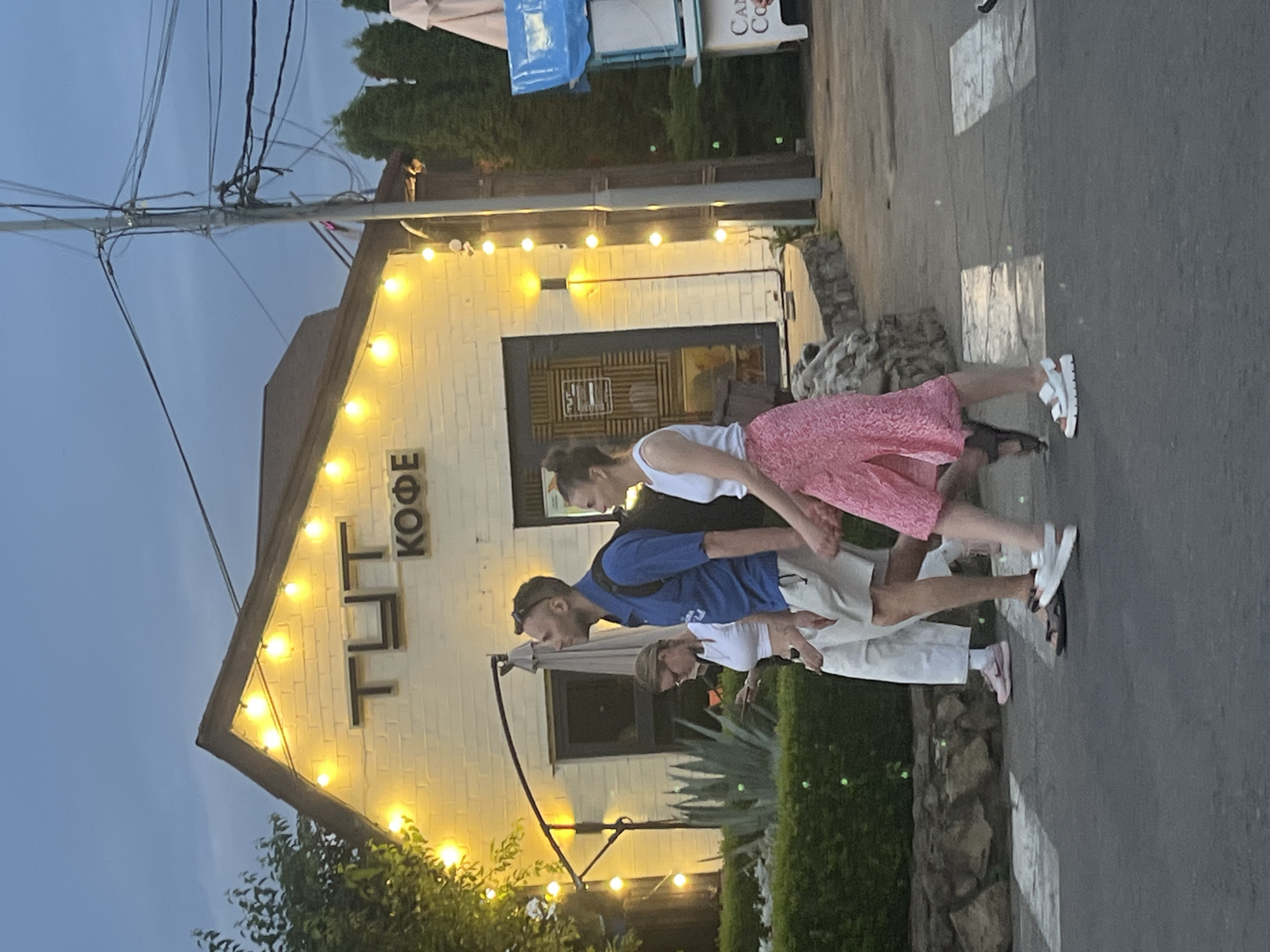 The image depicts a street scene in Krasnodar, Russia, with a building in the background featuring a sign that reads "ТУТ КОФЕ" (Here Coffee). The building is illuminated by a string of yellow lights. Three individuals are walking across a pedestrian crossing in the foreground. The person in the center, a man wearing a blue polo shirt and beige shorts, is looking to the right. To his left, a woman in a white crop top and light-colored pants is looking at her phone. To the right of the man, a woman in a white tank top and a pink floral skirt is walking and holding hands with the man. The sky is overcast, suggesting it is either dawn or dusk. In the background, to the right of the coffee shop, a vendor cart with a blue awning is visible, displaying the word "CAN" on its side.