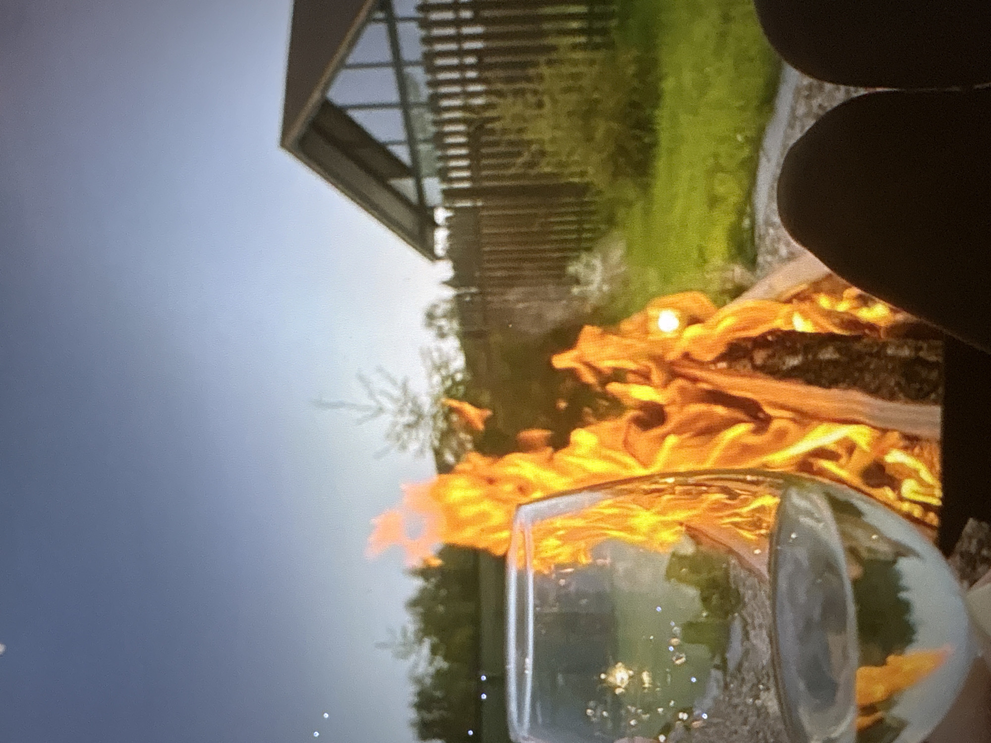 An outdoor campfire scene at dusk is depicted in this image. The foreground is dominated by a wine glass filled with a pale liquid, positioned in front of a brightly burning fire. The flames of the campfire are vibrant orange and yellow, casting a warm glow. Behind the fire, a wooden fence separates the grassy area from a modern-looking house with large windows and a dark roof. The sky above is a soft, gradient blue, transitioning to a lighter shade near the horizon. The overall atmosphere suggests a relaxed evening setting. The specific location in Pori, Finland is not identifiable from the image content.