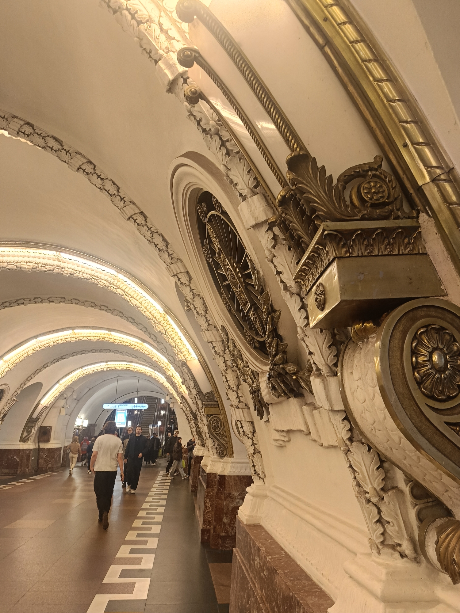 The image shows a corridor in the Avtovo Metro station in Saint Petersburg, Russia. The walls and ceiling are decorated with ornate white and gold baroque-style architectural elements, including arches, moldings, and decorative detailing. People are walking along the corridor, some heading towards escalators in the distance. The floor is tiled in brown with a geometric pattern. The image has a slightly warm, artificial lighting. The walls are marbleized at the bottom.
