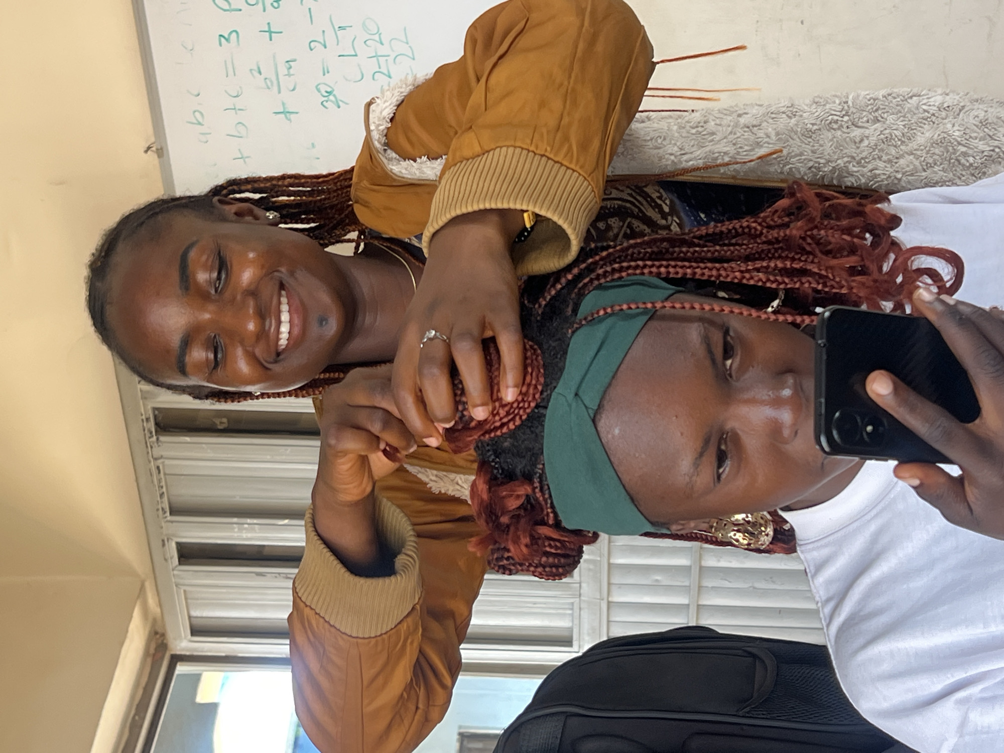 In Jos, Nigeria, a young Black woman with braided hair is having her hair styled by another young Black woman. The woman getting her hair done is wearing a green headwrap and a white t-shirt, and is looking down at her phone. The woman styling her hair is wearing a brown jacket and is smiling as she works on the braids. A whiteboard with mathematical equations is visible in the background.