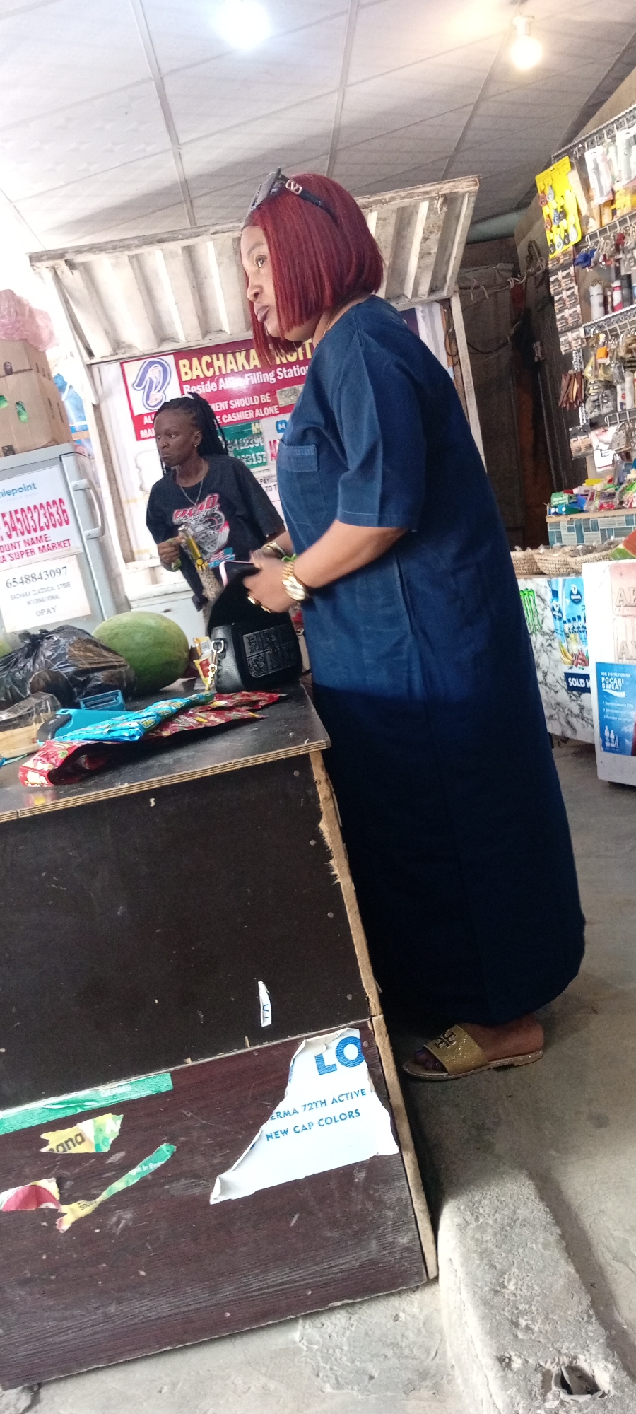 This media file captures an indoor scene at what appears to be a small market stall or shop, likely located in Aviation Quarters, Nigeria. The primary subjects are two individuals. In the foreground, a woman with striking red bobbed hair and sunglasses on her head is dressed in a long, dark blue outfit. She is looking to her right, her posture suggesting she might be interacting with someone or observing something off-camera. Beside her, a second person, seemingly a cashier or vendor, stands behind a counter, wearing a black t-shirt with a graphic print and looking towards the foreground.
The setting is a commercial space with merchandise visible. Shelves stocked with various goods, including what appear to be toiletries or small electronic accessories, are positioned in the background. The counter itself is cluttered with items such as plastic bags, a large green fruit (possibly a watermelon), and colorful wrappers.
The lighting suggests it could be daytime, though the enclosed nature of the stall makes it difficult to ascertain the exact time of day or weather conditions outside. The overall atmosphere appears casual and functional, typical of a local retail establishment.
Visible text in the image includes signage with brand names and contact information, such as "niepoint," a phone number "5450323636," and "BACHAKA NOTT Reside All Filling Station." Other text fragments like "MENT SHOULD BE E CASHIER ALONE" and "ERMA 72TH ACTIVE NEW CAP COLORS" provide further context about the services or products offered. The presence of these signs indicates a business operation in progress.