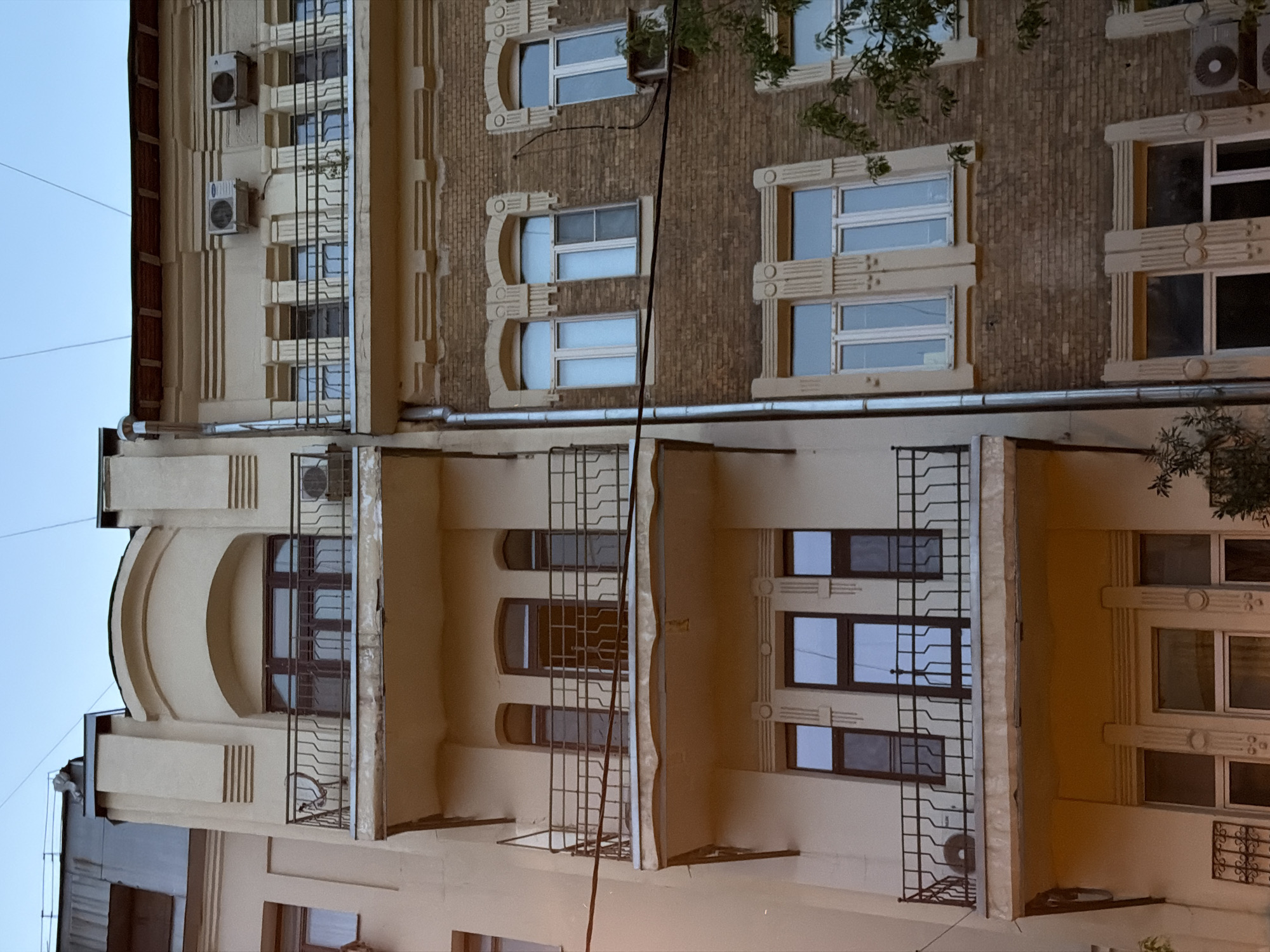 The image shows the facade of a building in Moscow, Russia, constructed from light brown bricks with symmetrical architectural elements. Windows are adorned with decorative frames and patterns. Air conditioning units are affixed to the building's exterior. Balconies with metal railings are present. Utility lines are visible extending along the building's surface. The lower portion of the image displays additional parts of the building, which include windows and balconies with similar railing features.