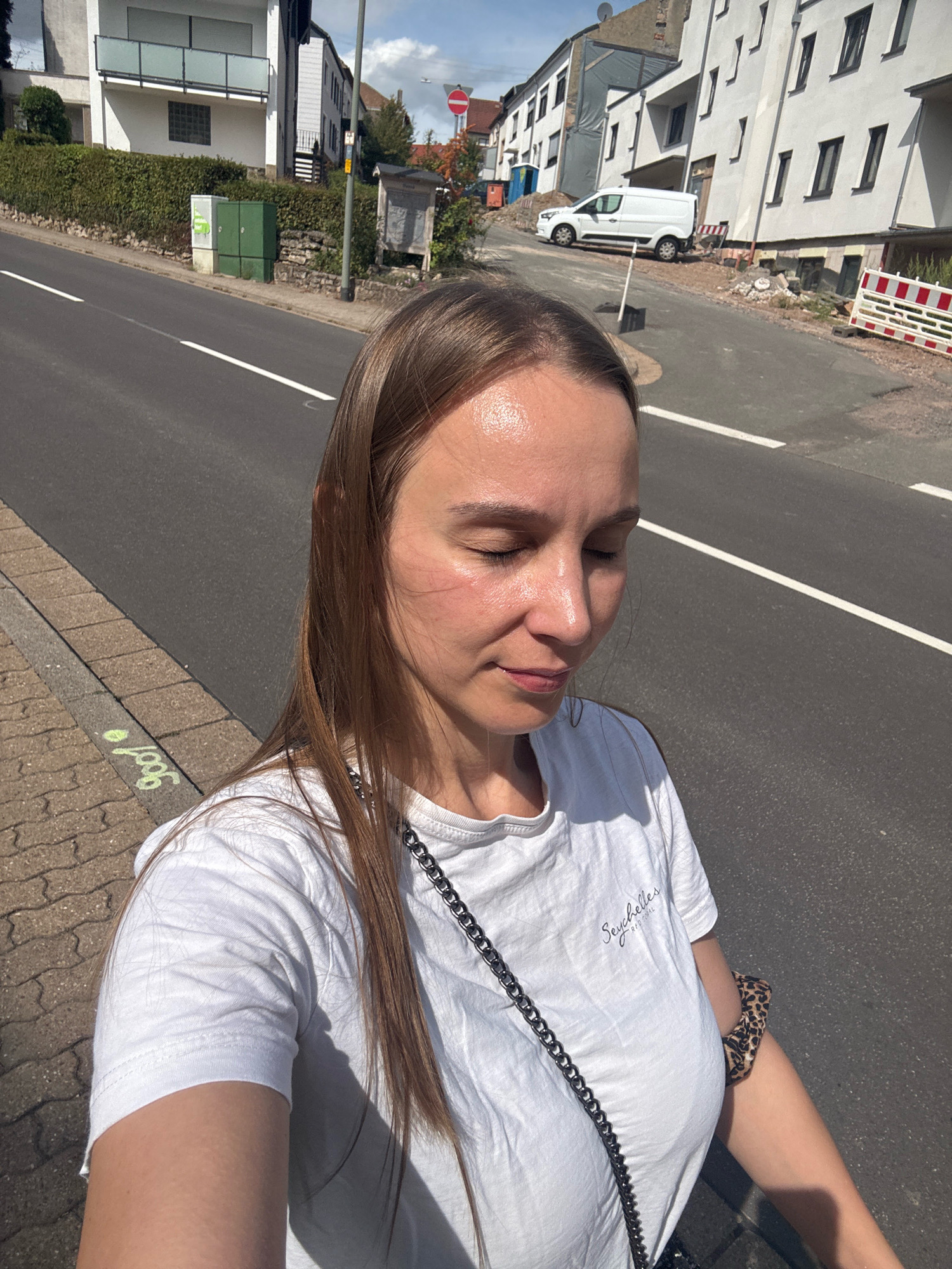 The image captures a young woman standing on a sidewalk next to a street in Überherrn, Germany. The woman is wearing a white t-shirt and has long brown hair. She is looking down with her eyes closed. The street is paved and has white lines marking the lanes. On the left side of the street, there is a sidewalk with a brick pattern, and further back, a green utility box is visible. Across the street, there are buildings, a white van, and construction barriers. The sky is cloudy.