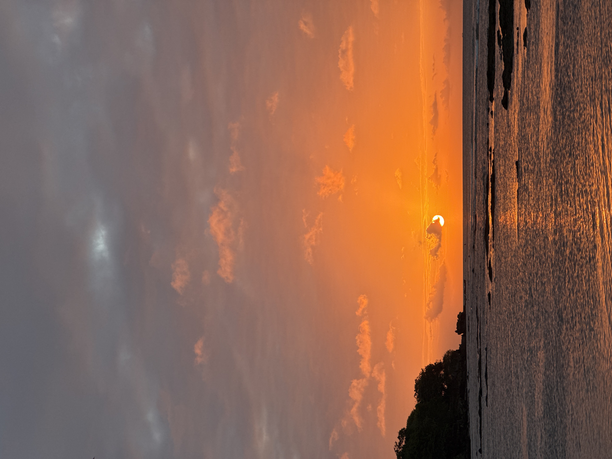 This is a photograph taken at sunset in Kuta Selatan, Indonesia. The image captures a dramatic sky with the sun low on the horizon, casting a warm, orange glow. The sky is a blend of soft grey clouds on the left, transitioning to vibrant oranges and yellows as they approach the sun. Wispy clouds are illuminated by the setting sun, creating a beautiful contrast against the deeper hues of the sky.
The sun itself is a bright, partially obscured disc, peeking out from behind a small cloud. Rays of light emanate from it, fanning out across the sky.
The foreground of the image features the ocean, with the water reflecting the colors of the sunset. The surface of the water appears calm, with gentle ripples catching the light. Along the right edge of the frame, there is a dark silhouette of land, likely a coastline or a small island covered in dense vegetation, which is also bathed in the warm sunset light.
The overall mood of the photograph is serene and awe-inspiring, characteristic of a tropical sunset. The time of day is clearly evening, as indicated by the setting sun and the warm, diffused light. There is no visible text in the image.