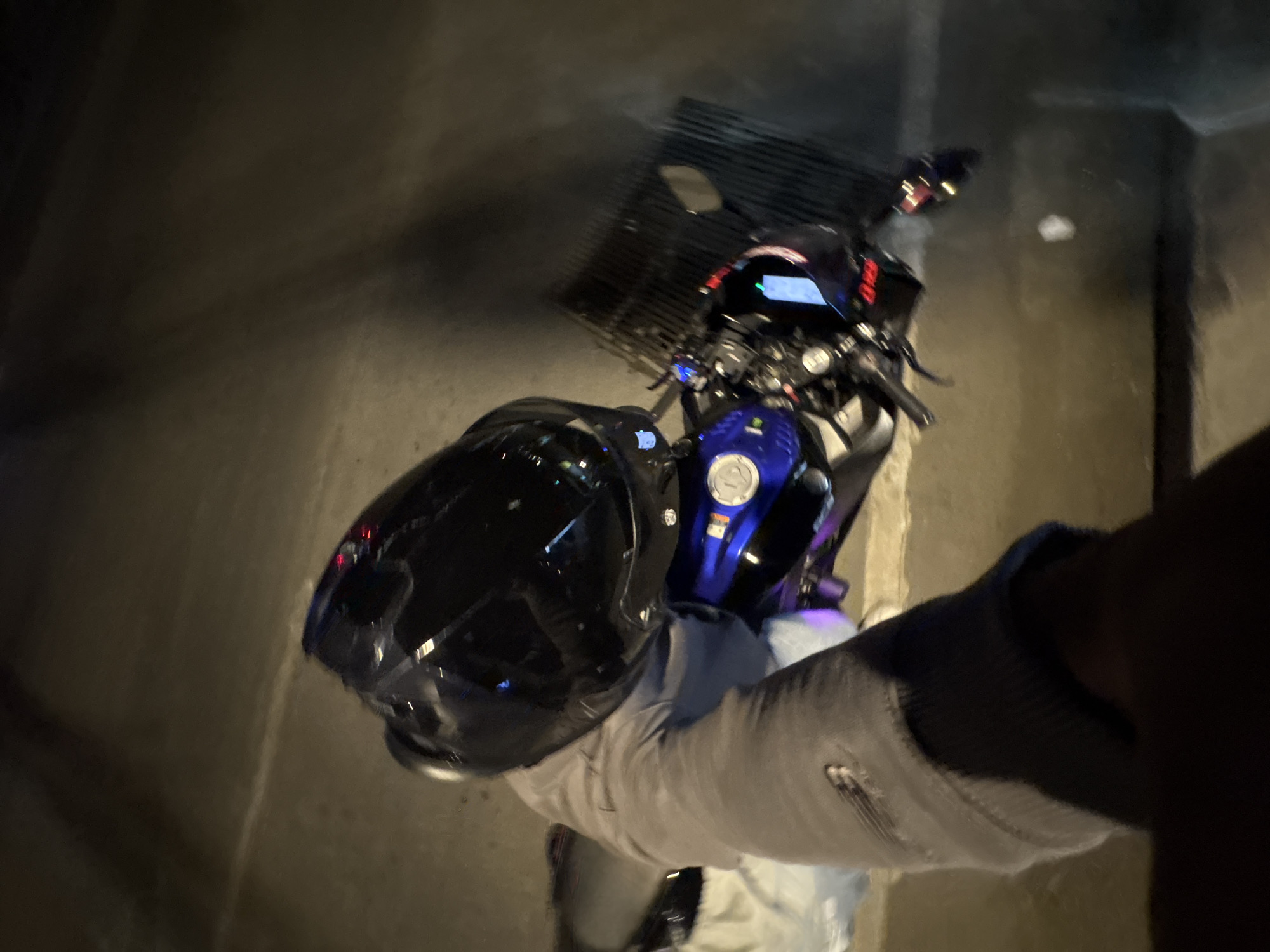 The image depicts a low-light, overhead view of an individual and a motorcycle in Davao City, Philippines. A person, whose lower body from mid-thigh down and a portion of a forearm are visible, is positioned on or immediately adjacent to a motorcycle. The individual is wearing grey pants with a zippered pocket detail on the upper right thigh, and a dark sleeve covers the visible forearm. A black, full-face motorcycle helmet rests securely on the person's right thigh, featuring a reflective visor. The motorcycle is a blue and black sportbike or naked bike, with its blue fuel tank, silver fuel cap, handlebars, front brake lever, and a digital instrument cluster displaying red illuminated numbers clearly discernible. A rearview mirror extends from the right side of the motorcycle. The ground surface is dark, likely concrete or asphalt, and a dark metal grate, consistent with a storm drain, is visible to the right of the motorcycle. The scene is illuminated by artificial light sources, indicating nighttime conditions.