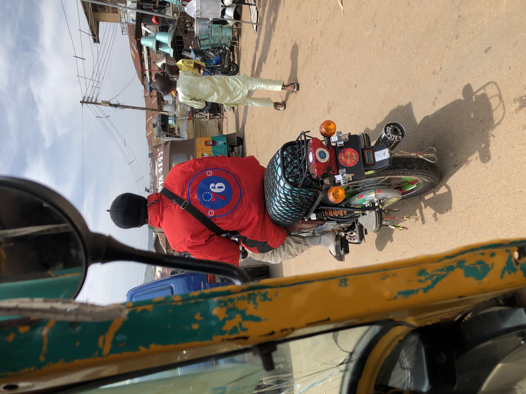A rider wearing a red jacket and a black beanie is on a motorcycle in an outdoor marketplace. The motorcycle has a black mesh cargo carrier and a red taillight assembly with orange turn signals. The rider's jacket has a circular emblem with the number "6" and a starburst design. In the background, there are buildings with corrugated metal roofs, utility poles with many wires, and other vehicles, including a three-wheeled auto-rickshaw. Two young men in traditional light-colored clothing are walking in the middle ground. The scene suggests a busy commercial area in Jos, Nigeria, characterized by its lively street life and distinctive architecture.