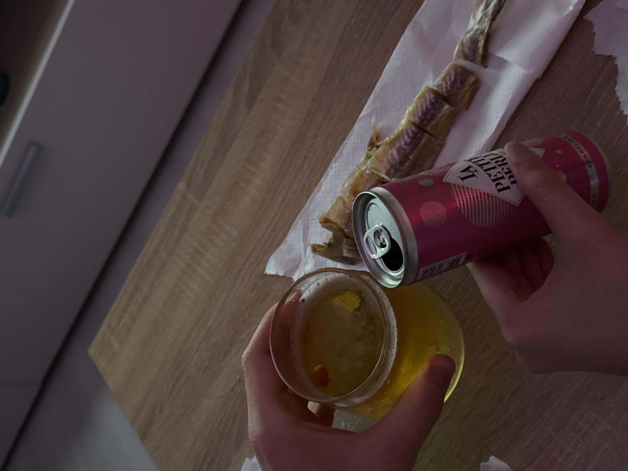 A high-angle shot depicts a partially consumed glass of yellow liquid, with a pink and white can labeled "LA PETIT DERU" being poured into it by a pair of hands. The scene is set on a wooden surface, which also features a piece of white paper on which sits a sliced fish product. The location is Unknown, Unknown.