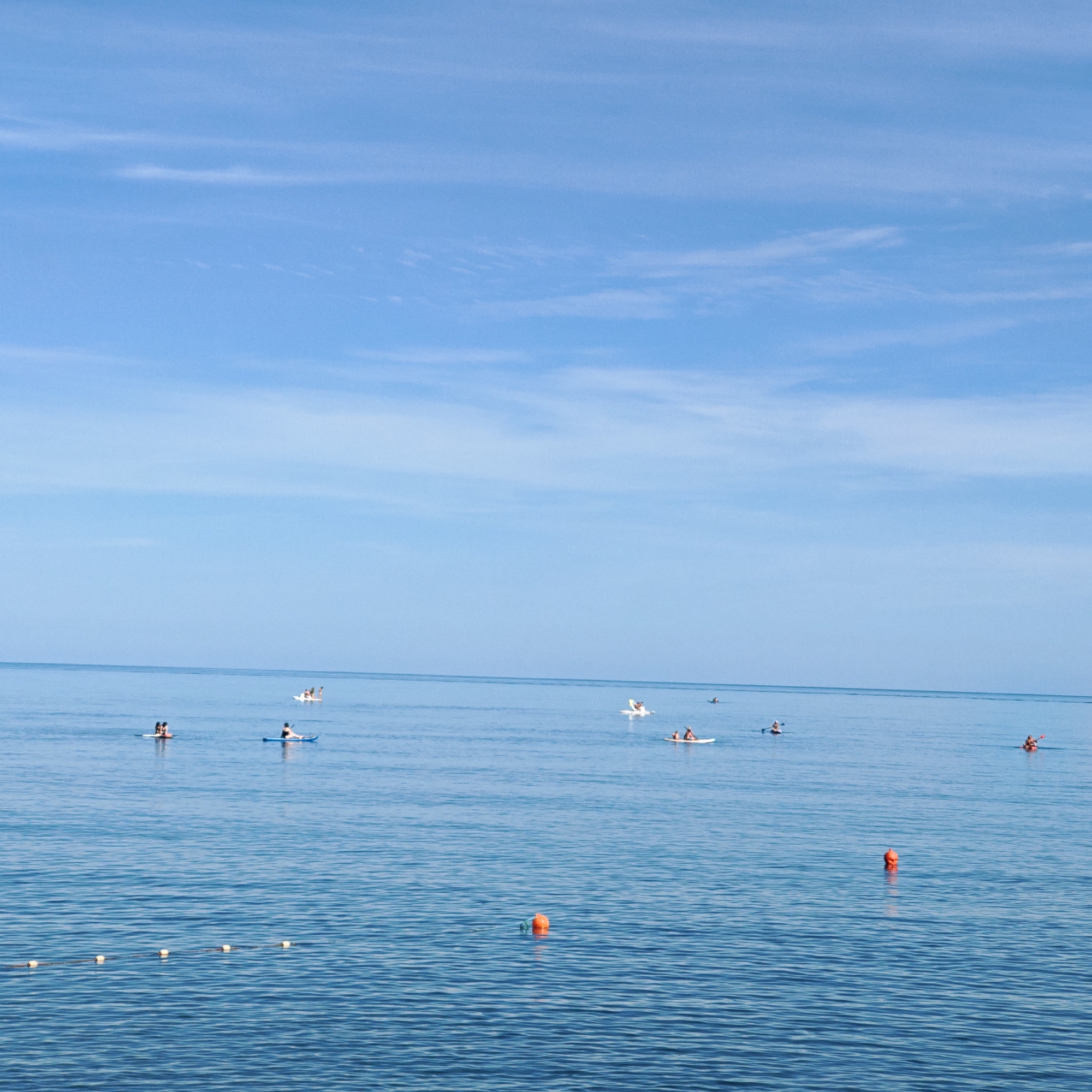 The image depicts a serene seascape under a clear blue sky with wispy white clouds. The vast expanse of calm, blue water dominates the frame, with a gentle horizon line separating it from the sky. Scattered across the water are several people engaged in various water activities.
In the foreground, a rope with several buoys floats on the surface, marking a boundary in the water. Further out, multiple individuals are seen on paddleboards and kayaks. One group of three is on a paddleboard, while another person is on a kayak to their right. Further in the distance, a small group of people are on what appears to be a floating structure or a larger paddleboard. To the far right, another individual is kayaking. In the middle distance, there are a few more paddleboarders scattered across the water.
The scene suggests a leisurely day at the beach or on a lake during daylight hours, likely late morning or afternoon given the bright sky. The weather appears to be pleasant and calm, suitable for water sports. The overall mood is peaceful and relaxed, with the individuals enjoying the water and the open expanse. No visible text is present in the image.