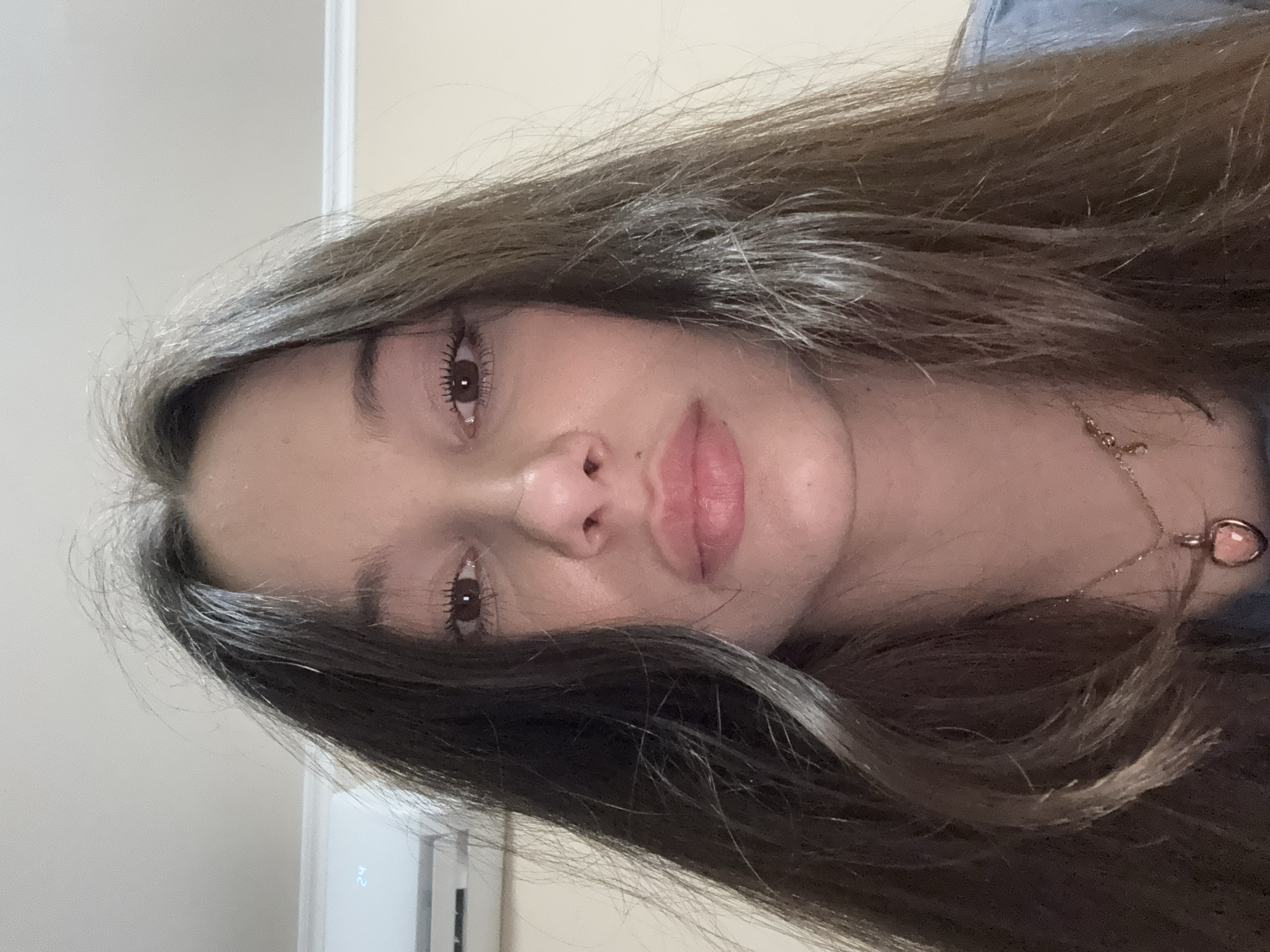 A close-up, low-angle, indoor selfie of a young woman with long, dark brown, wavy hair. She is looking directly at the camera with a neutral expression. Her eyes are brown, and she is wearing mascara. She has full lips with a subtle pink tint. She is wearing a delicate gold necklace with a heart-shaped pendant. The background is a plain, light-colored wall, and a white air conditioning unit is visible on the left side of the frame, displaying the number "24". The lighting is soft, and the overall scene appears to be in a residential setting. There is no indication of the specific location within the city of Sofia, Bulgaria.