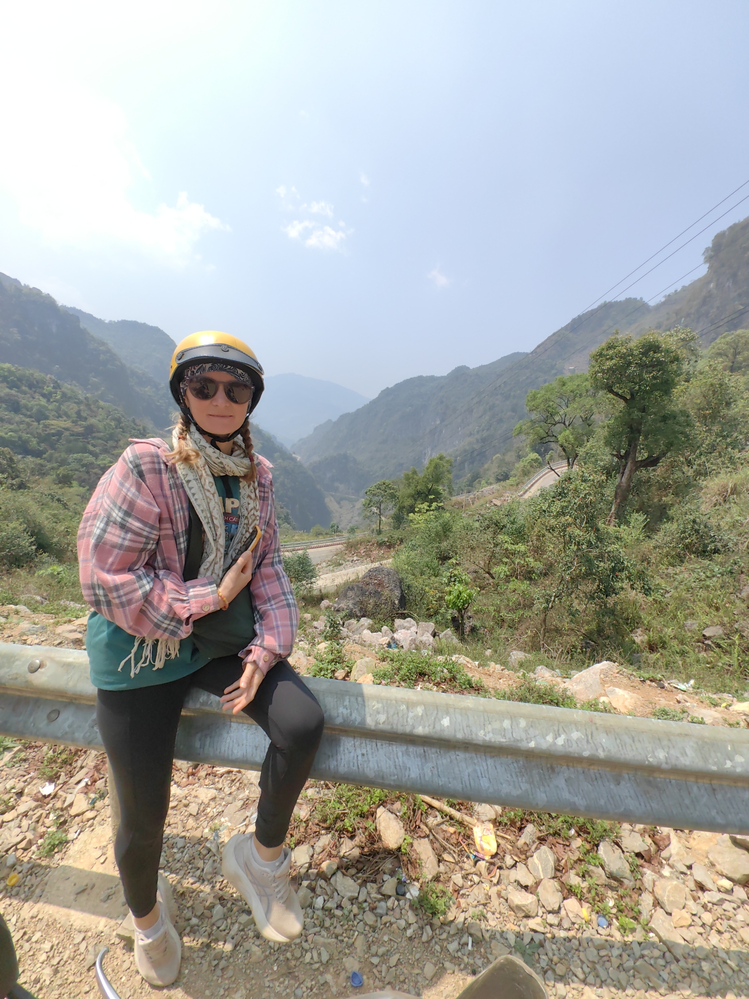 The image depicts a woman wearing a yellow helmet, sunglasses, a pink and white plaid shirt, and black leggings. She is leaning against a grey metal guardrail. Her hair is in braids. In the background, there is a mountainous landscape with green vegetation and a winding road. The ground is covered in rocks and dirt. The location is Xã Xuân Trường, Vietnam.