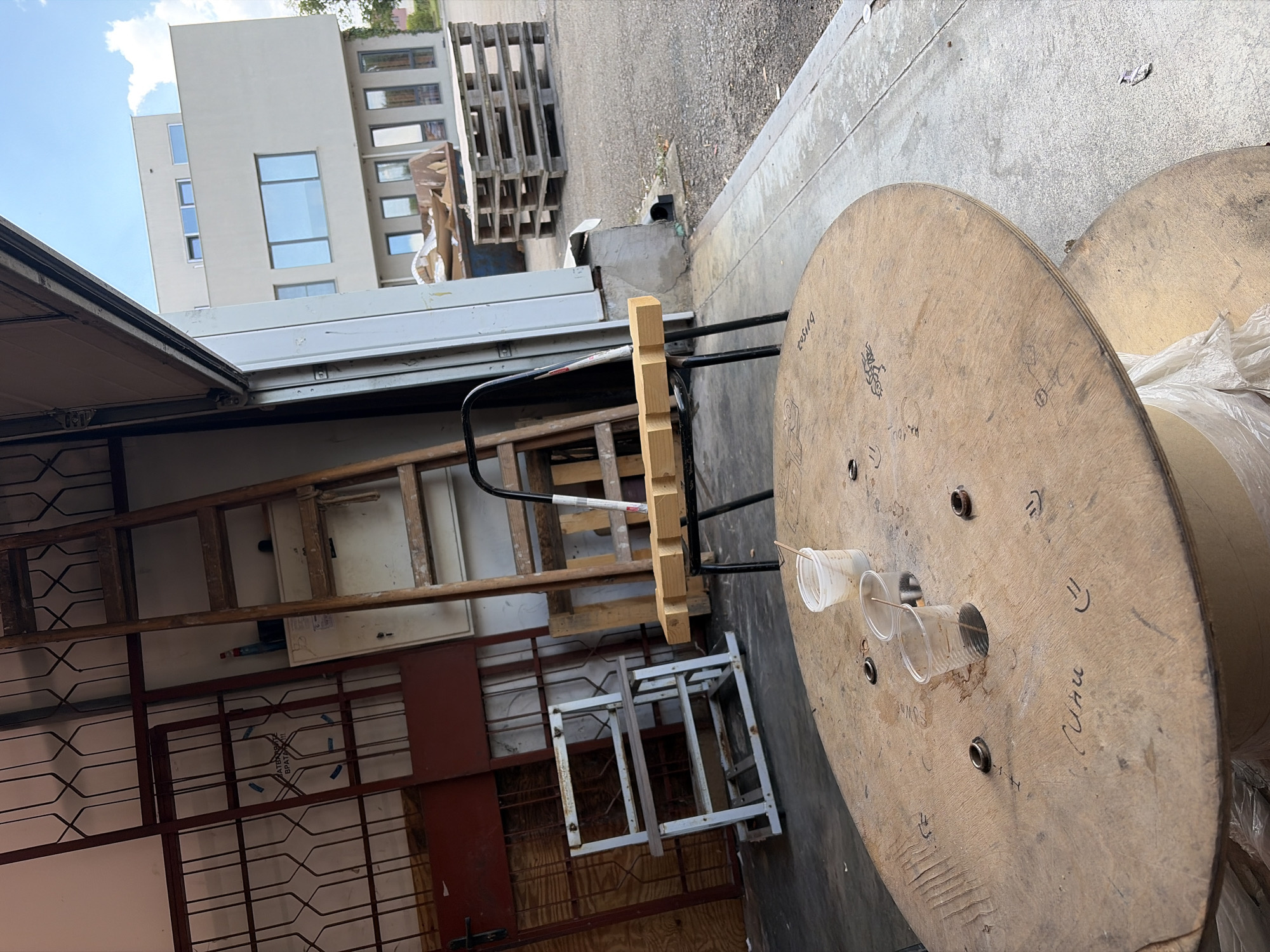 This is an outdoor scene in Ravno pole, Bulgaria, likely a workshop or storage area. In the foreground, a large, round wooden spool is positioned on the ground. Two clear plastic cups, each containing a liquid and a stick, are placed on the spool. The spool itself has various markings and some ring-shaped metal objects embedded in it. Behind the spool, a metal chair with a wooden plank across its seat is visible. To the left, a tall wooden ladder leans against a wall, partially obscuring some metal grates and red-colored panels. Further in the background, a modern, multi-story building with large windows stands under a blue sky with scattered clouds. A large dumpster filled with debris and a stack of wooden pallets are also present in the background, suggesting ongoing construction or industrial activity. The ground is paved with concrete and gravel.