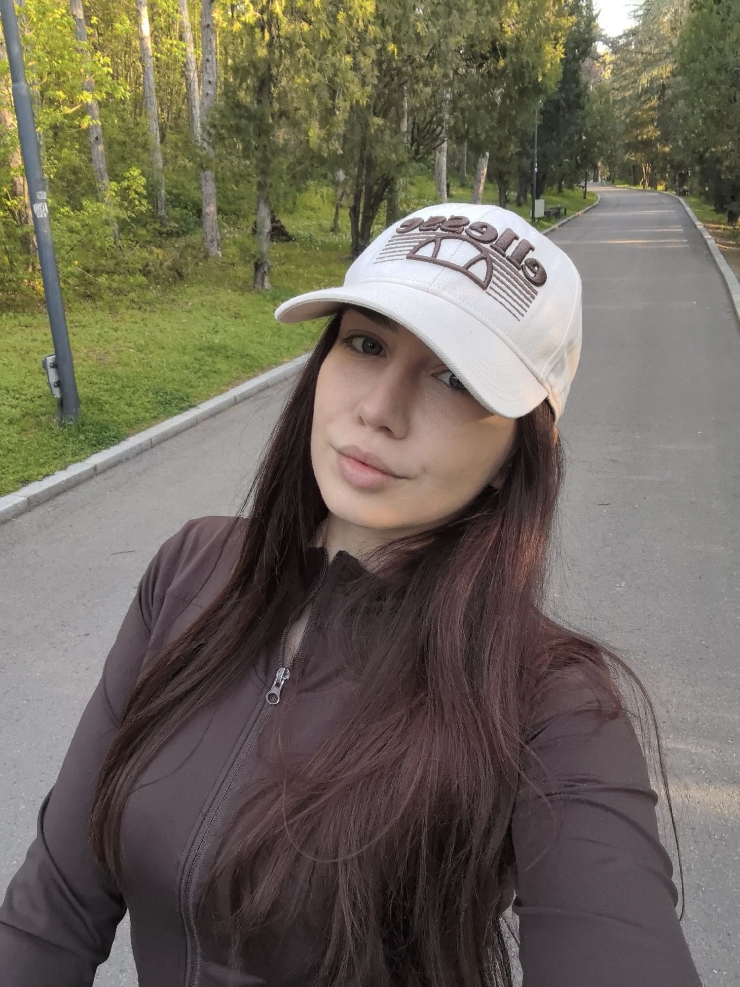 This image captures a selfie of a young woman enjoying an outdoor setting, likely a park or a tree-lined avenue in Stara Zagora, Bulgaria. The woman is the primary subject, positioned centrally, looking directly at the camera with a serene and slightly smiling expression. She has long, dark brown hair flowing over her shoulders and is wearing a light-colored, possibly off-white, baseball cap. The cap features a prominent dark brown embroidered logo which, due to the selfie perspective, appears mirrored, spelling "esse||e" with its characteristic semi-circular emblem, identifying it as the Ellesse brand. She is dressed in a dark brown, long-sleeved jacket or top with a front zipper.
The background depicts a pleasant natural environment. A wide, paved path, suitable for walking or jogging, stretches into the distance, bordered on both sides by vibrant green grass and numerous mature trees. The path gently curves to the right, implying a longer route through the park. The lighting suggests late afternoon or early evening, often referred to as "golden hour," as warm, soft sunlight illuminates the scene, particularly the tops of the trees, and casts gentle shadows. The weather appears clear and agreeable. On the left side of the frame, a dark grey utility pole is partially visible. The overall scene evokes a sense of calm and leisure in a beautiful outdoor space.