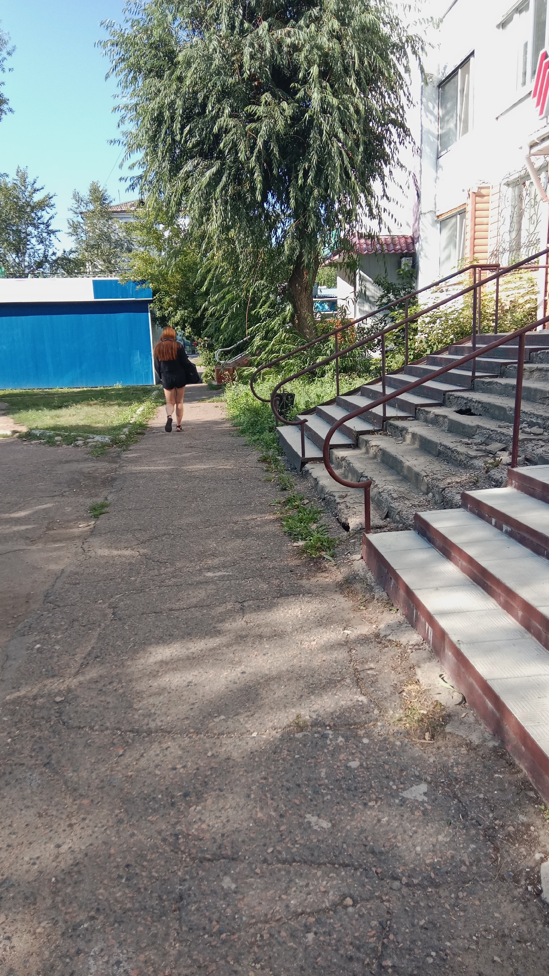 A woman with long, auburn hair is walking away from the camera on a concrete pathway. She is wearing a dark, loose-fitting jacket or coat and dark shorts. To her right, a set of concrete stairs with metal railings ascends alongside a building. The building is light-colored with windows. To her left is a bright blue structure. A large tree with drooping branches shades a portion of the scene. The environment appears to be outdoors, possibly in a residential area or an institutional complex in Petropavl, Kazakhstan, on a sunny day. The concrete pathway shows signs of wear and tear, with cracks and patches of grass growing through.