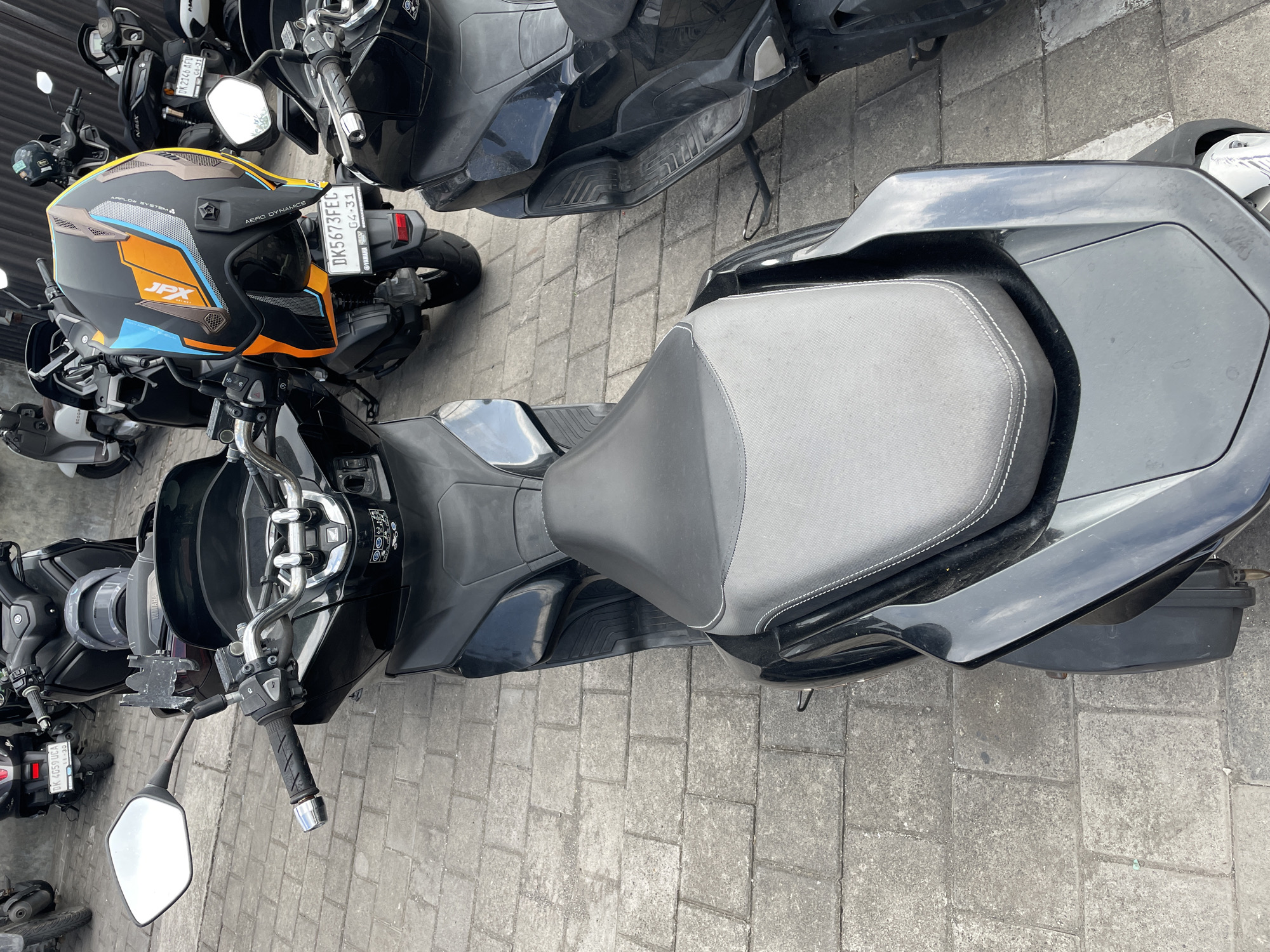This is an eye-level, medium shot of several parked motorcycles and scooters on a paved surface in Mengwi, Indonesia. The foreground is dominated by the seat of a black scooter, with white stitching visible on its textured surface. To the left of this scooter, a silver rearview mirror is attached to the handlebars. A black motorcycle helmet with orange, blue, and brown accents is resting on the handlebars of a scooter behind the foreground scooter. Its license plate reads "DK5673FEC". Other motorcycles and scooters are visible in the background, parked closely together. The ground is made of light gray, rectangular pavers. The scene appears to be an outdoor parking area.