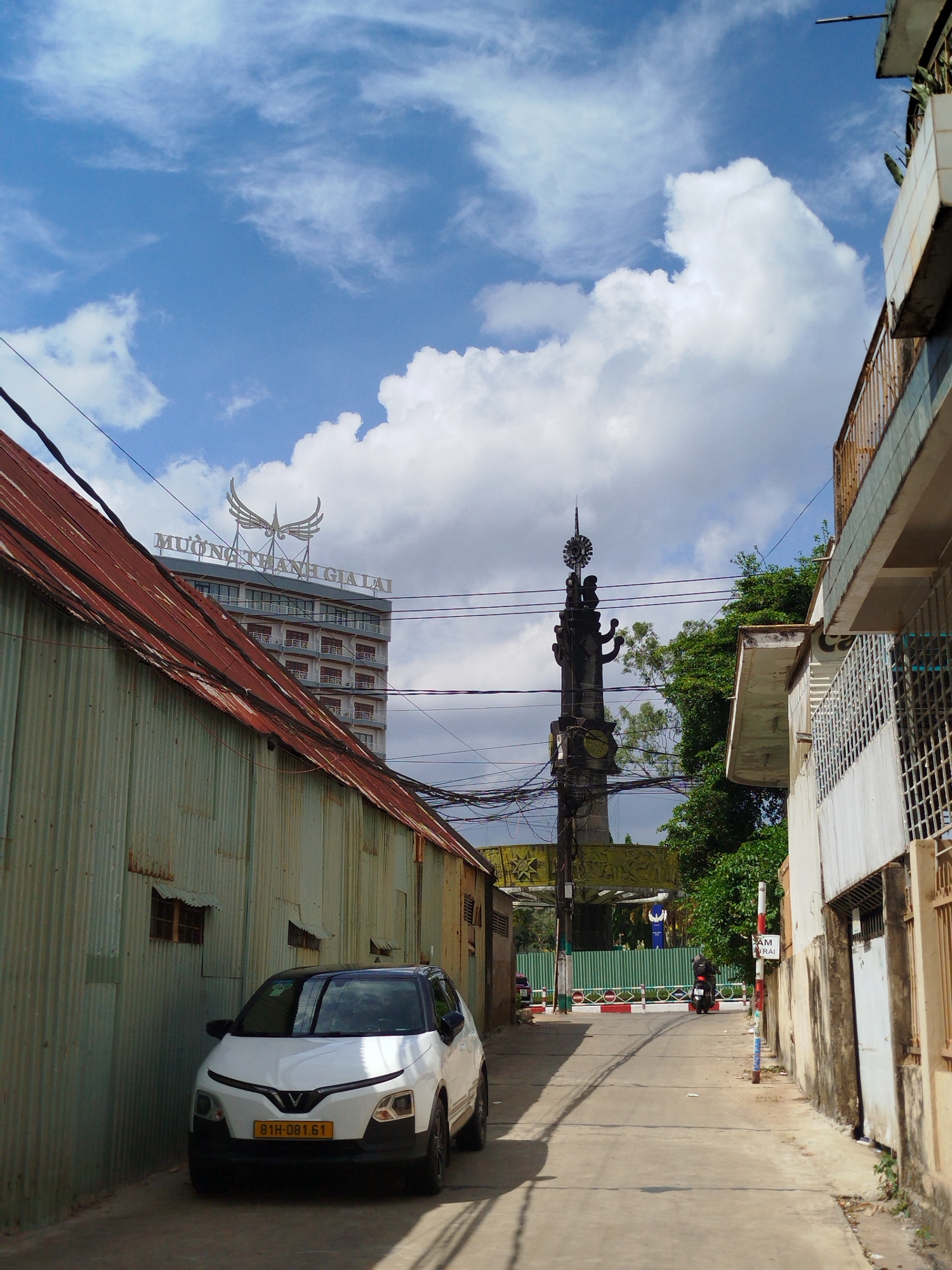 A daytime scene shows a narrow street in Pleiku Ward, Vietnam. The street is flanked by buildings, one corrugated metal, the other a multi-story building with balconies. A white car is parked along the corrugated metal building. In the distance, a monument topped with a sunburst-like design is visible. Utility wires crisscross above. A building with the sign "MUONG THANH GIA LAI" is partially visible behind the corrugated metal building. A person on a motorcycle is further down the street, near a striped barrier. The sky is blue with scattered clouds.
