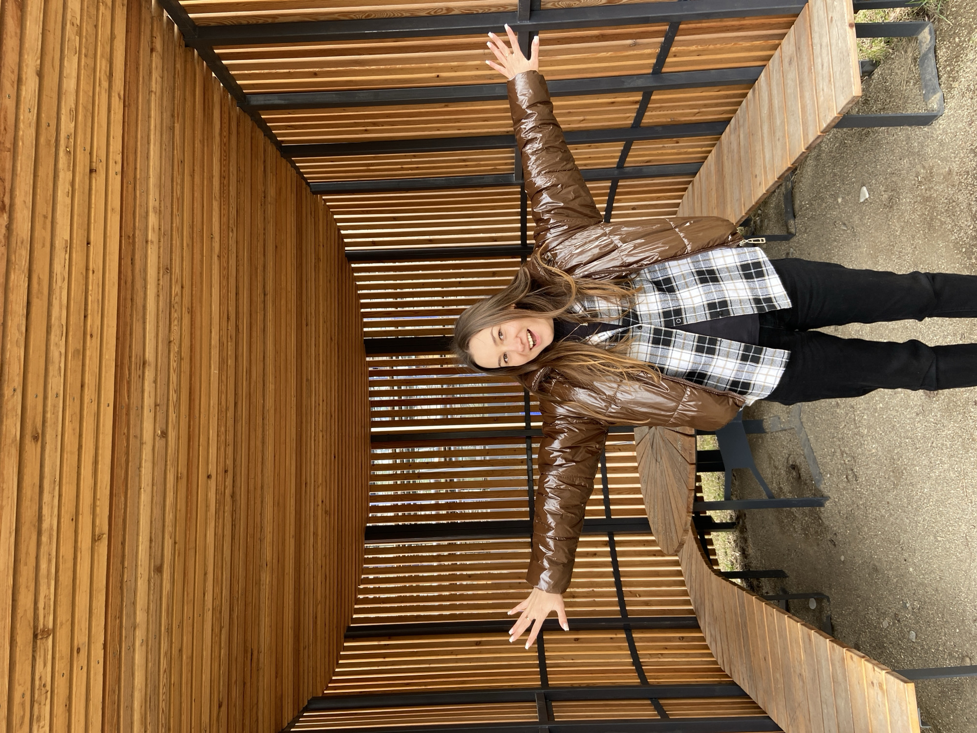 A young woman is standing inside a wooden structure, possibly a gazebo or pavilion, in what appears to be a park or outdoor recreational area. The structure consists of vertical wooden slats forming walls and a roof made of horizontal wooden slats spaced apart to allow light to filter through. The interior also contains curved wooden benches. The woman has her arms outstretched to her sides, indicating a gesture of openness or joy. She is wearing a shiny brown puffer jacket over a black and white plaid shirt, black pants, and dark footwear. Her hair is long and light brown. The ground outside the structure is a mix of dirt and gravel.
While I can describe the scene in detail, I cannot definitively confirm the location as Izhevsk, Russia without additional information or visual cues specific to that city.