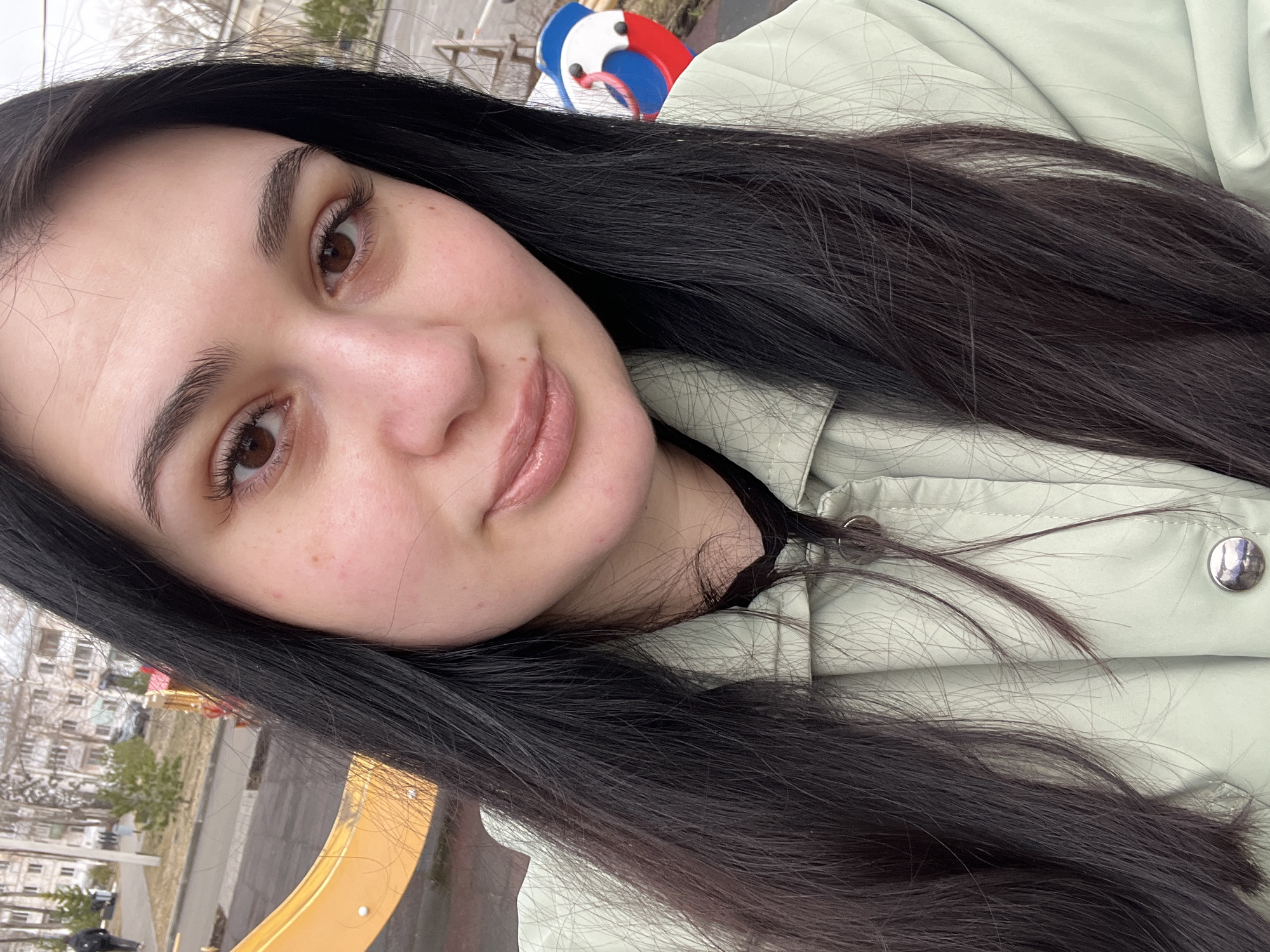 A woman with dark hair and light brown eyes is taking a selfie. She is wearing a light green jacket. In the background, there is a playground with a yellow slide and a red, white, and blue structure. Beyond the playground, there are buildings and trees, suggesting an urban environment. This location is in Chaikovsky, Russia.