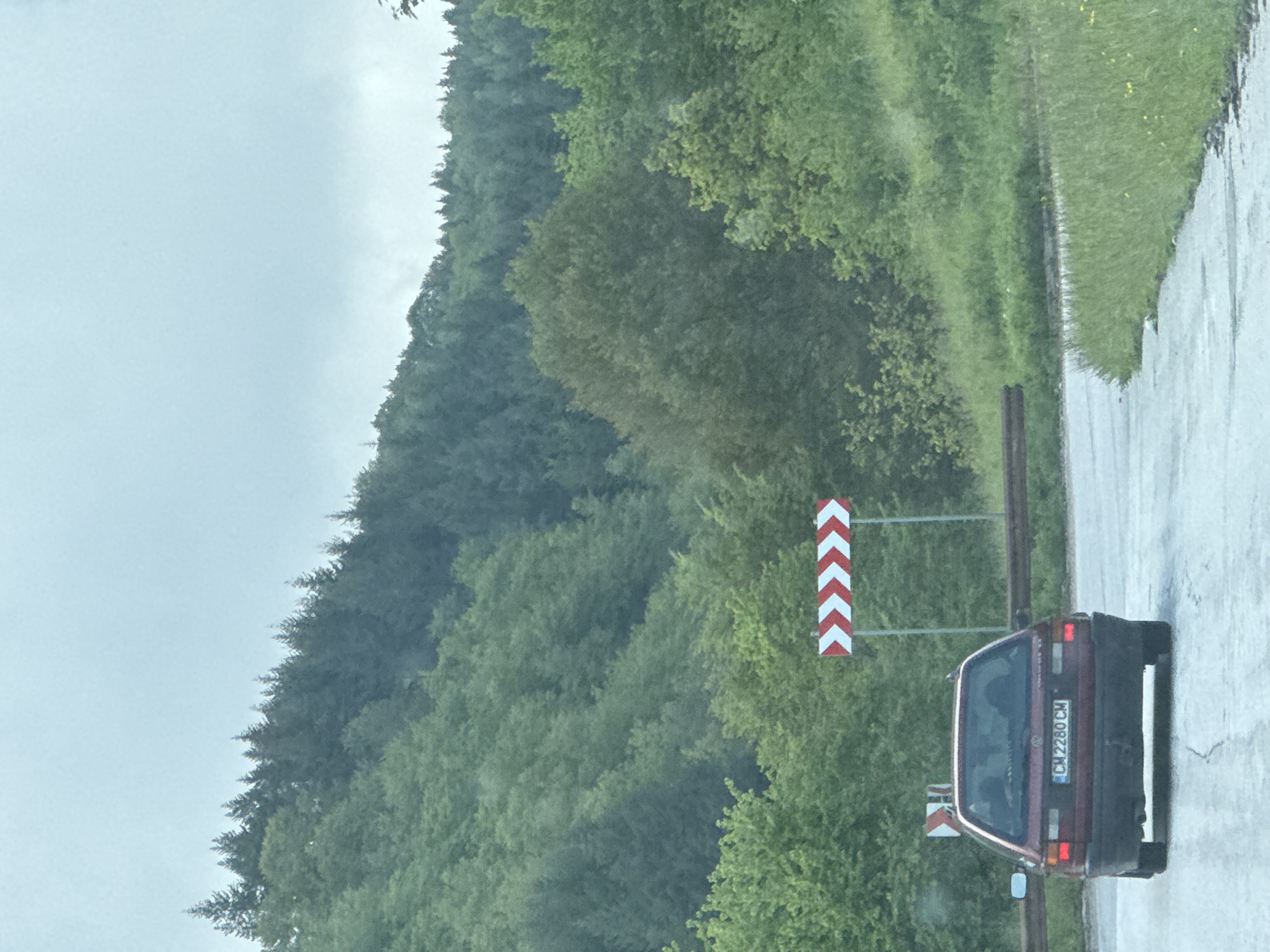 The image depicts a road in a rural area, likely in Chokmanovo, Bulgaria. A maroon Volkswagen Golf is parked on the road. A red and white chevron road sign is next to the vehicle, indicating a sharp bend or curve in the road. The license plate of the car reads "CM 2280 CM". The road surface appears wet, suggesting recent rain. The background consists of a forested hillside covered with dense trees and vegetation. The sky is overcast, indicating cloudy weather. The overall scene is one of a rural roadside with a parked car.