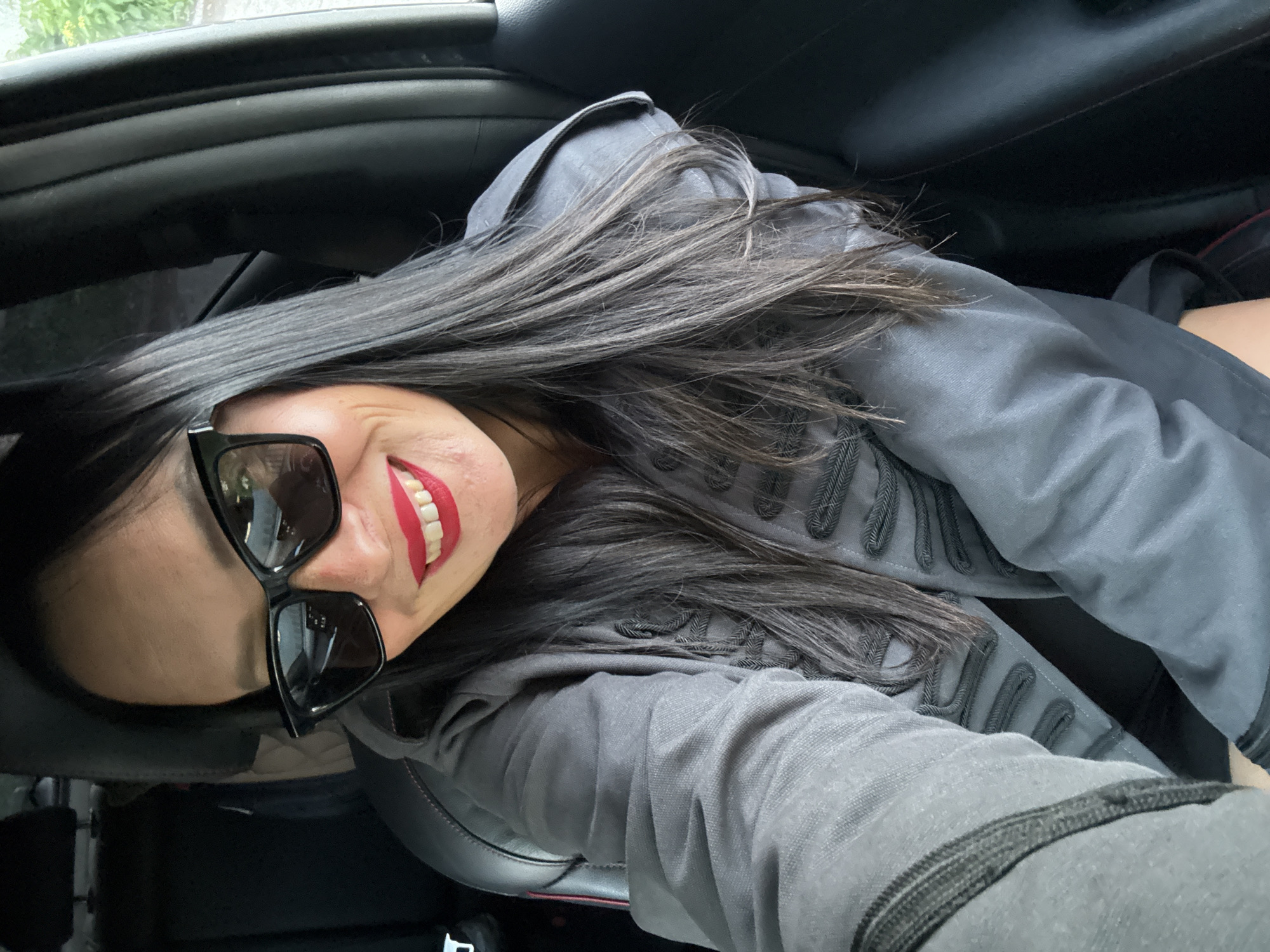 A woman with long, dark hair, wearing sunglasses and red lipstick, is in the driver's seat of a car. She is smiling and looking at the camera. The car appears to be parked. The background is blurred, showing some greenery outside the car window. The image is a selfie taken by the woman. The location is not specified in the image.