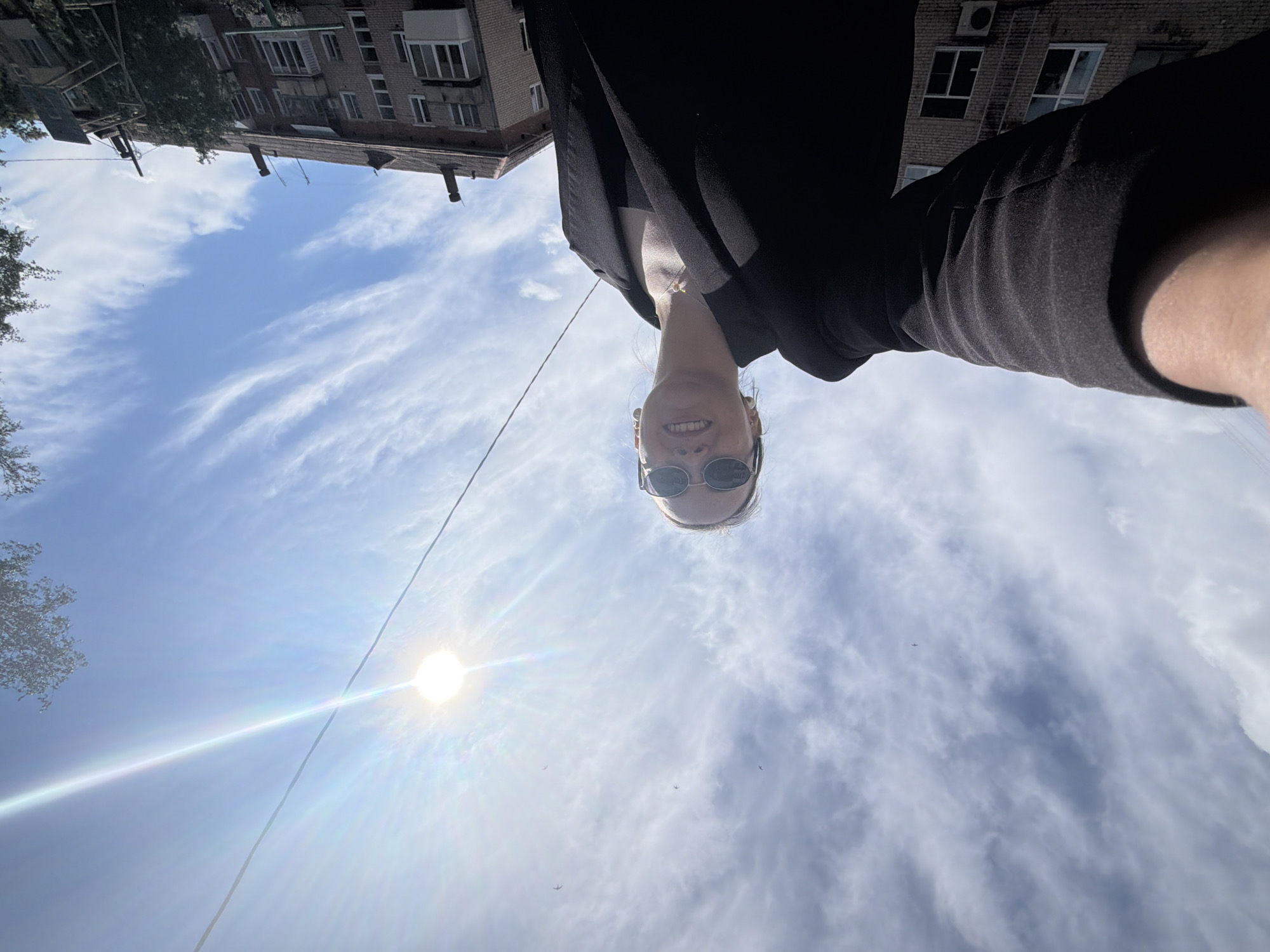 A low-angle selfie is taken by a young woman wearing sunglasses and a black blazer. She is smiling and looking upwards towards the sun, which is bright and centered in the sky, creating lens flare. Wispy clouds are visible against a bright blue sky. In the background, apartment buildings constructed of brick are visible, characteristic of urban architecture in Chaikovsky, Russia. The scene suggests a sunny day outdoors in a residential area of the city.