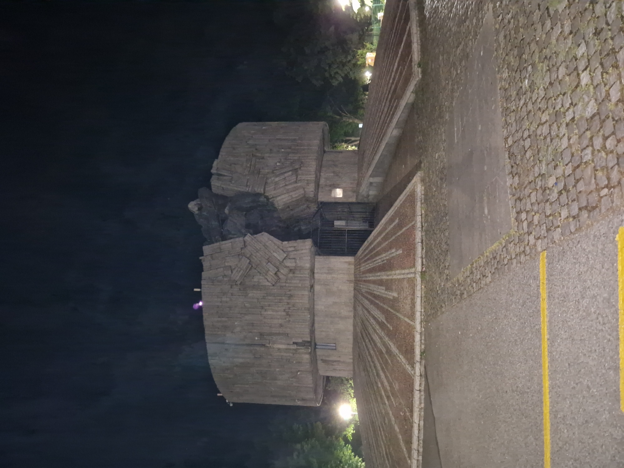 The image captures a nighttime scene dominated by a large, multi-tiered concrete or stone structure next to a dark body of water. The structure, possibly a monument or a unique building, features a blocky, angular design with various levels, appearing to be composed of stacked and interlocking elements. A bright street lamp illuminates the area near its base on the right, and smaller lights are embedded within the structure itself. A metal gate or railing is visible at a lower entrance point of the structure.
To the right of the main structure, the ground transitions from a patterned section with radiating lines of brick or stone to an asphalt or concrete surface. This paved area displays two distinct yellow lines, suggesting a designated parking space or road. Further to the right, a textured cobblestone path, partially overgrown with moss or weeds, extends across the foreground.
The left side of the image is largely occupied by a dark, reflective body of water, likely a river or lake, which mirrors some distant light sources. In the background, dark silhouettes of trees and foliage are discernible against the dim night sky, dotted with a few additional lights. The overall atmosphere is serene and dark, with artificial lighting accentuating architectural details and pathways. No specific activity is taking place, and no text is visible in the frame.
