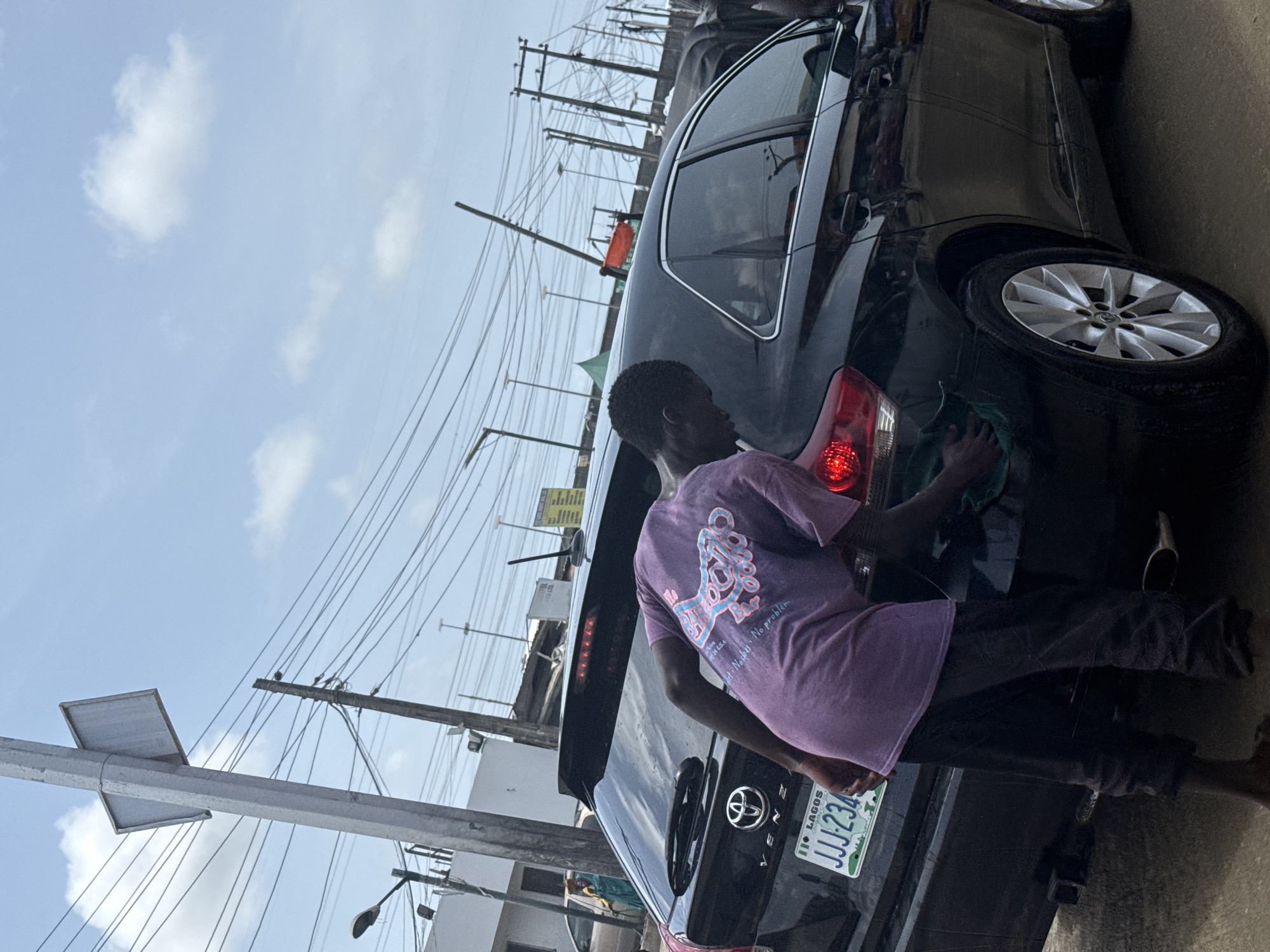 A man is washing the back of a black car. The license plate reads "JJJ 234", indicating Lagos, Nigeria. The location appears to be an outdoor setting in the city of Ado, Nigeria, as the image shows utility poles, wires, and a cloudy sky. The man is wearing a purple shirt and dark pants, and he is using a cloth to clean the car's taillight. The environment suggests an urban or semi-urban area, with elements of infrastructure such as poles and wires.