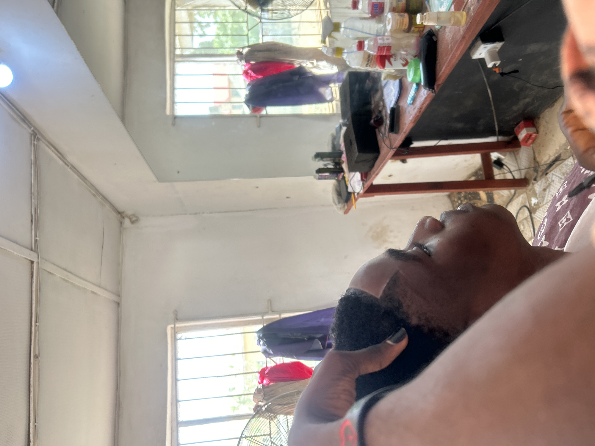 A man is getting a haircut at an unknown location. The room has white walls and a window with a fan in front of it. There is a table with various bottles and objects on it. The man getting the haircut is African American with short hair. The person giving the haircut is also African American and is holding clippers. The location is in the city of Unknown, Unknown.