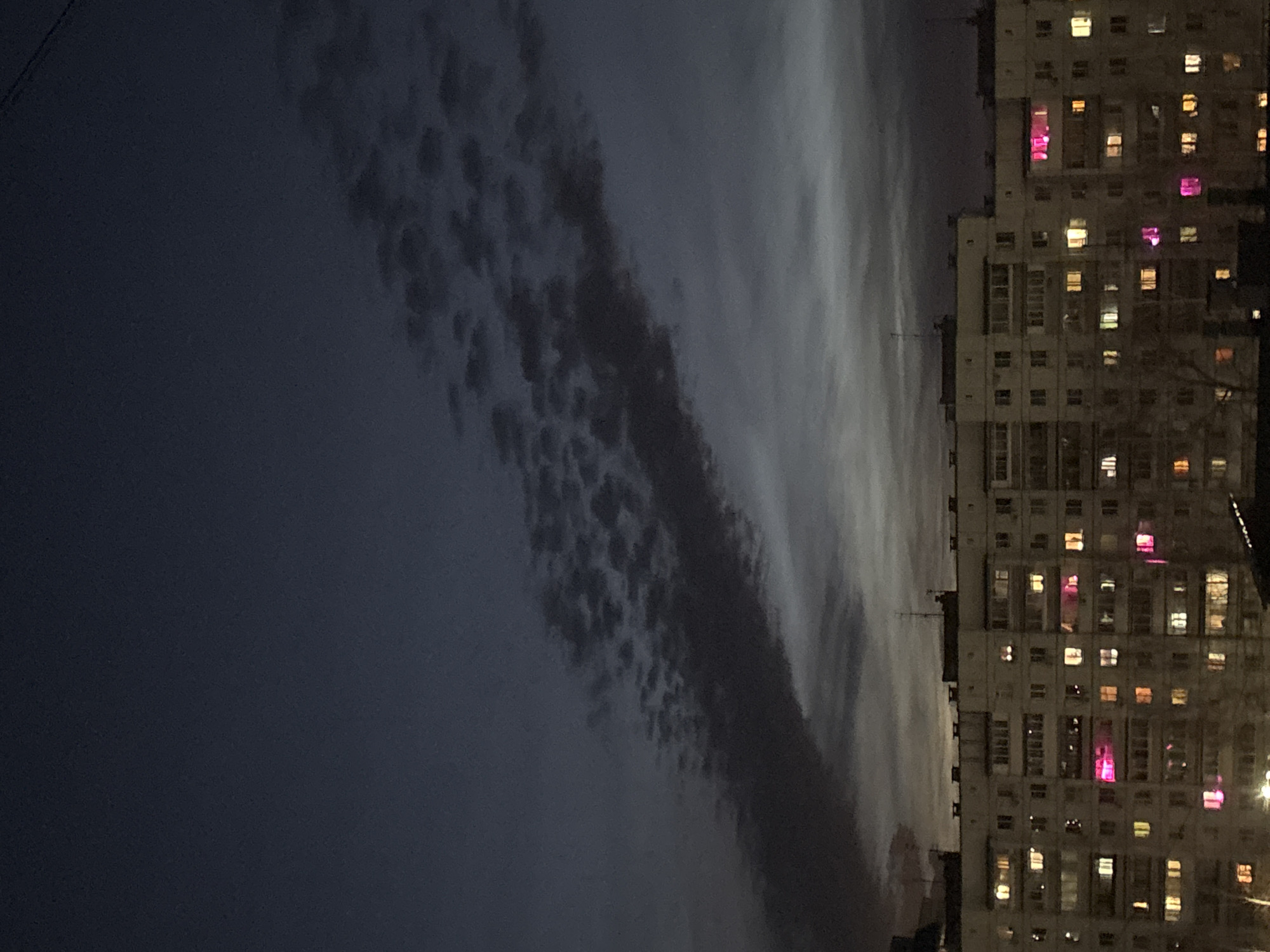 The image depicts a nighttime scene in Chaikovsky, Russia, showcasing an apartment building illuminated with lights. Several windows emit a pinkish glow. The sky above is dark, with a stripe of cloudy formations, creating a contrast with the building. The buildings are dense and towering, indicating an urban residential area. There is no sign of people visible.