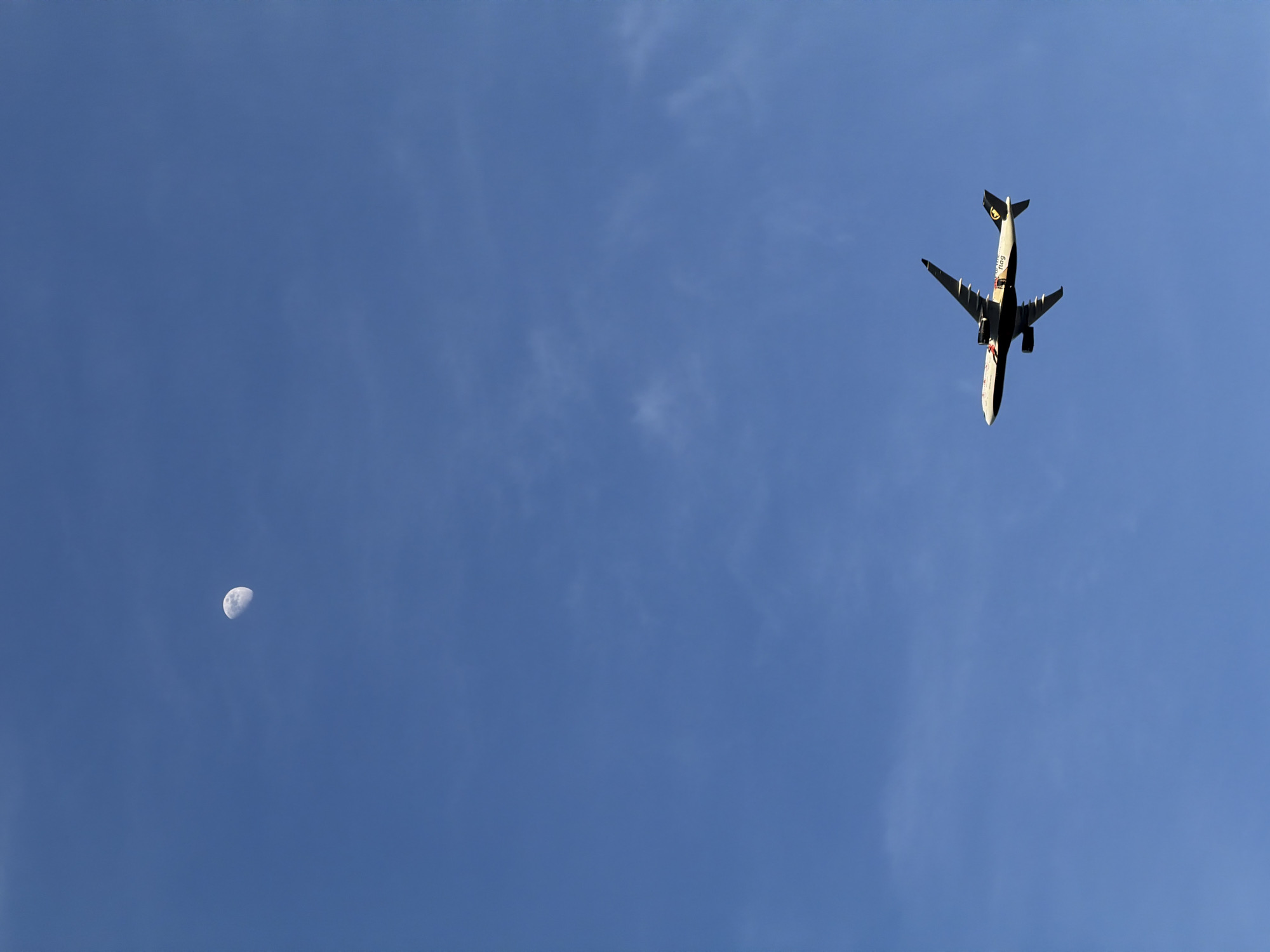 The image shows a blue sky with a half-moon in the lower-left corner and an airplane flying overhead on the right. The underside of the airplane is visible. No location in Montreal, Canada is discernible from the image.