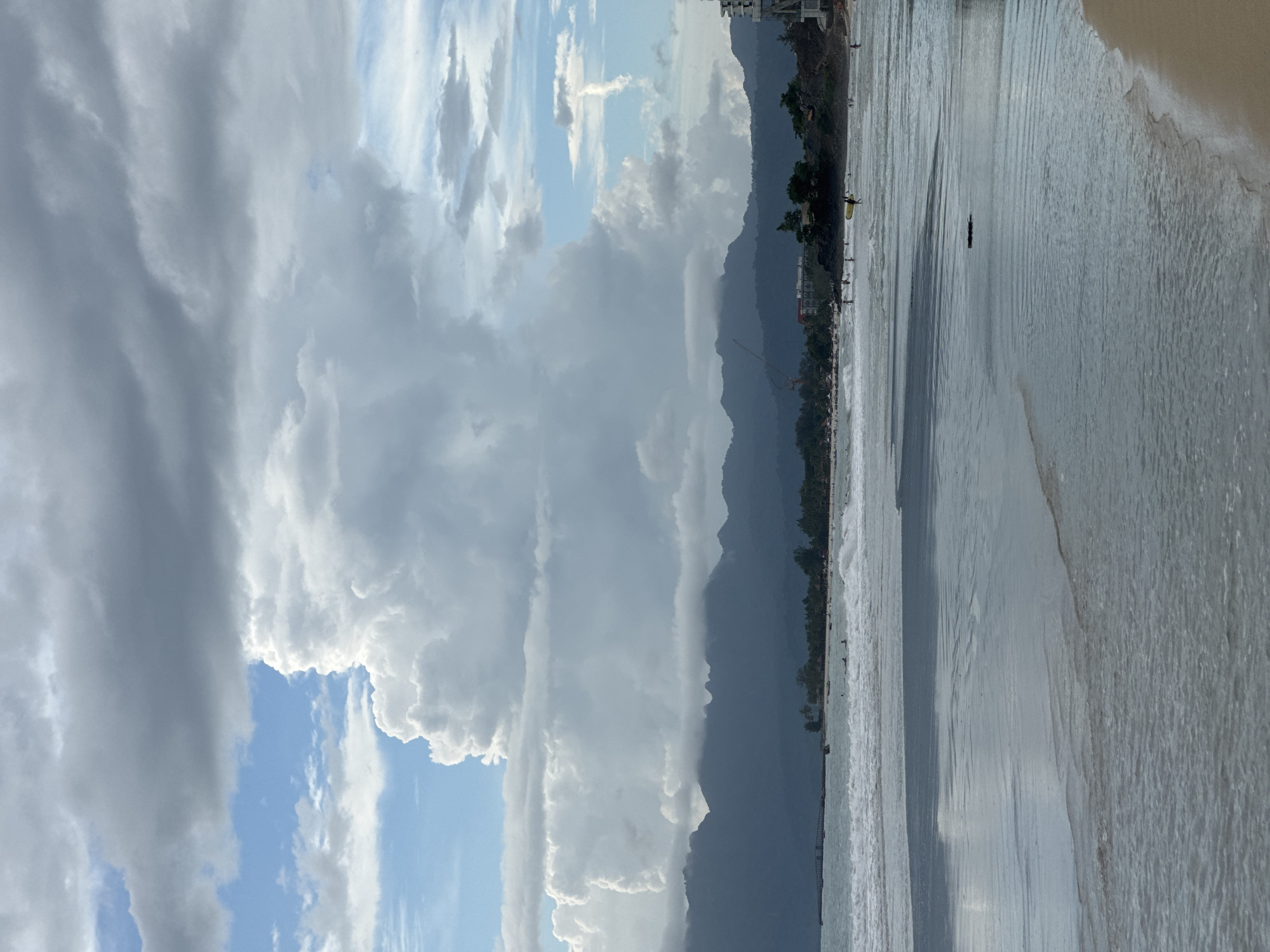 A wide shot captures the serene coastal landscape of Tengah Island, Malaysia, with the ocean dominating the foreground. Gentle waves lap the sandy shore, reflecting the dramatic sky above. Dark, silhouetted mountains form a jagged horizon in the distance, their peaks softened by a layer of clouds. The sky is a tapestry of contrasting clouds, with large, fluffy white formations rising majestically against patches of bright blue. These cumulus clouds cast shadows, creating a dynamic play of light and dark across the scene.
A stretch of beach, dotted with various individuals, runs along the base of the mountains. Some people are visible in the water, seemingly engaged in recreational activities like swimming or surfing. A few buildings and palm trees can be seen along the coastline, hinting at a resort or inhabited area. The overall atmosphere is one of tranquility and natural beauty, with the imposing mountains and the vast expanse of the sky creating a sense of awe.