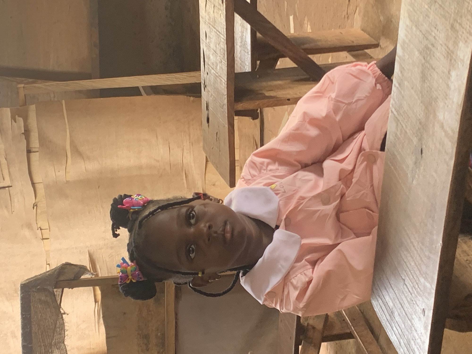 A young Black girl with braided hair and colorful hair clips, wearing a pink collared jacket, sits among rough wooden structures, likely makeshift furniture or shelves. The setting appears to be a simple interior space, possibly a school or home in Ukwu-Abwa, Nigeria, judging by the image context. The environment is rustic, with unadorned surfaces. The girl looks directly at the camera.
